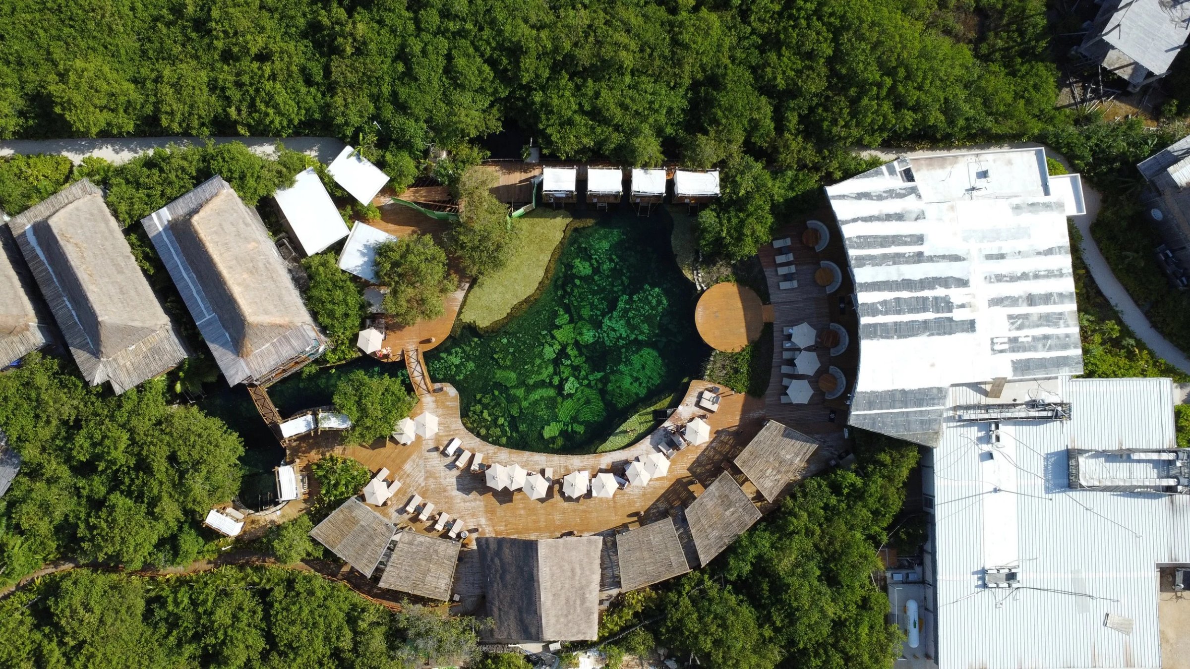 Aerial view of a resort with a central swimming pool surrounded by wooden decks, lounge chairs, and umbrellas, with thatched roof buildings and green trees surrounding the area.