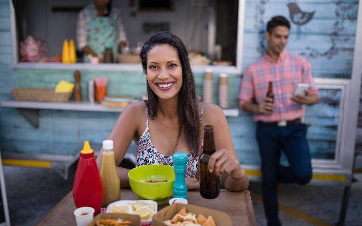 A happy looking woman posing for the camera as she is about to enjoy a Caribbean meal with a beer in her hand. The backdrop features a local eatery with the server preparing food and another person holding a drink in their hand