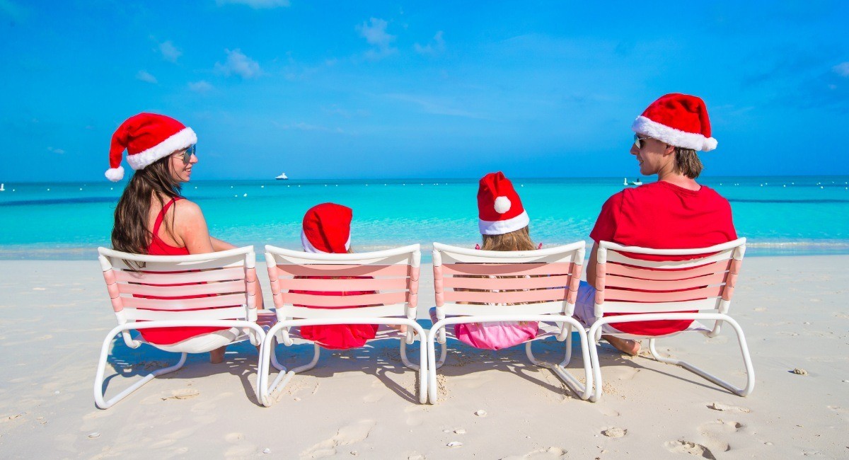 Family of four wearing Santa hats sitting on beach chairs overlooking turquoise Caribbean Sea during Grand Cayman Christmas vacation