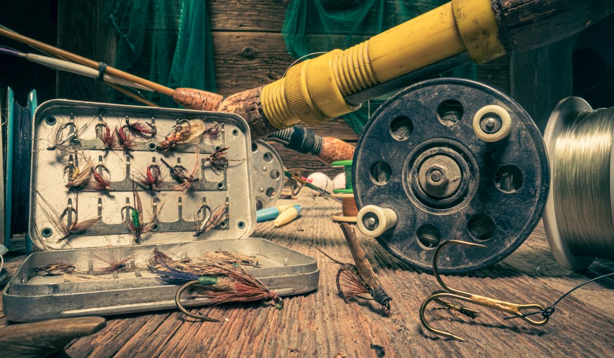 A set of fly-fishing accessories on display in a shed with an assortment of fly in a box, a fishing reel and some hooks.