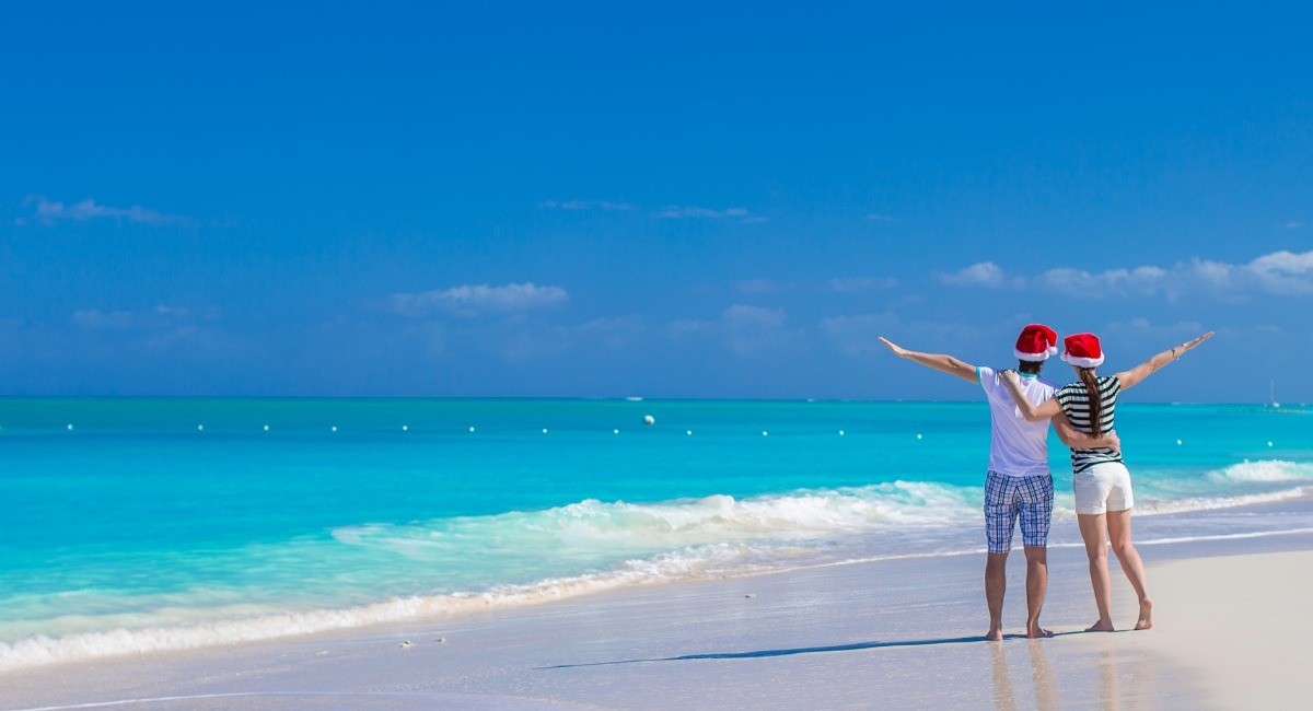 Happy couple wearing Santa hats celebrating a romantic Christmas on pristine Seven Mile Beach Grand Cayman with arms raised in joy