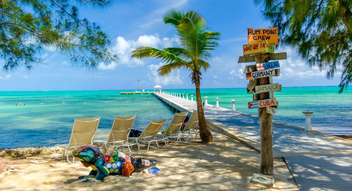 Rum Point Grand Cayman beach with wooden dock, lounge chairs, palm tree and directional distance signs showing destinations like Germany, Jamaica and Cuba