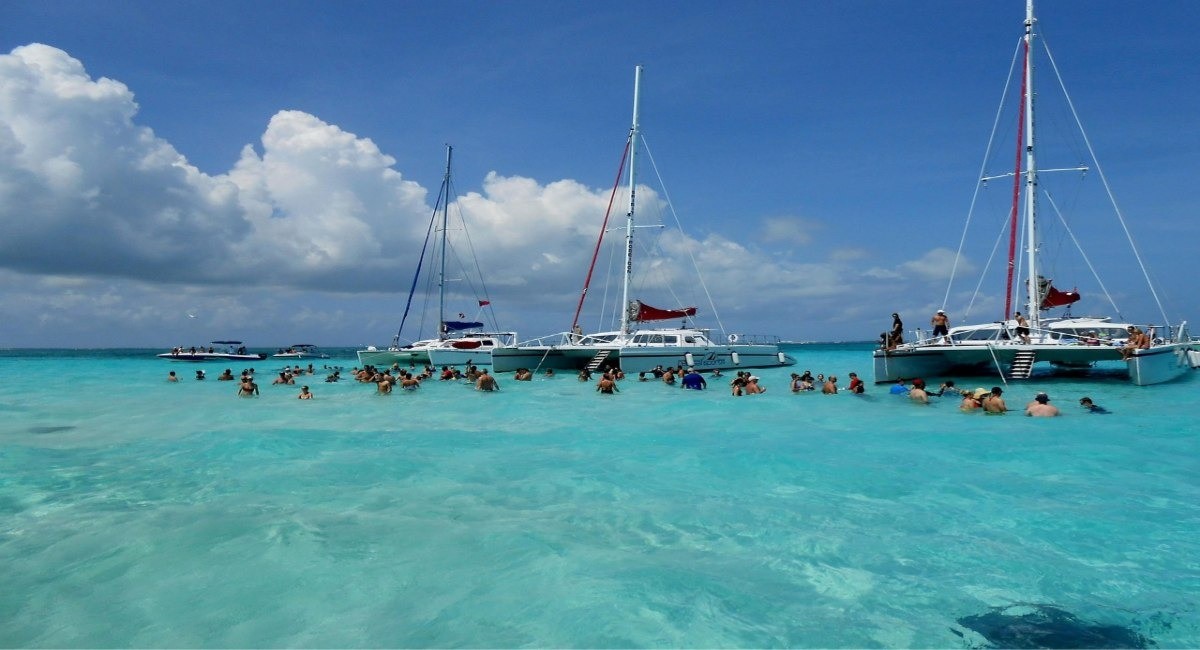 Tourists swimming in shallow turquoise water at Stingray City, Grand Cayman with multiple tour boats anchored nearby