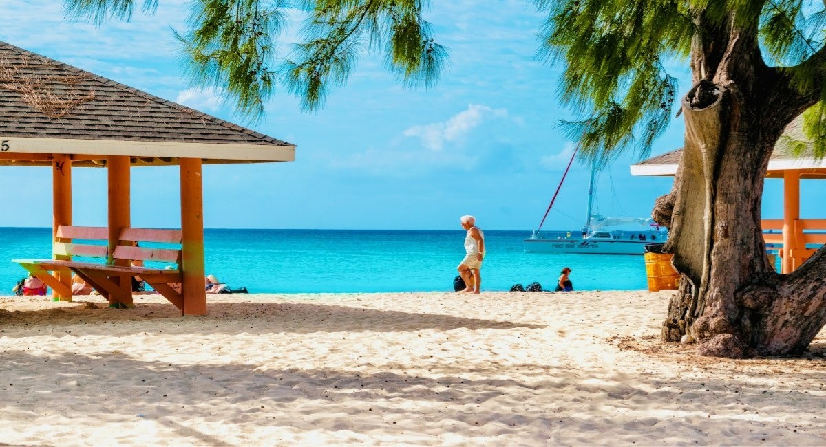 Seven Mile Beach with shaded beach pavilions, families relaxing on white sand, and a catamaran anchored in calm turquoise waters