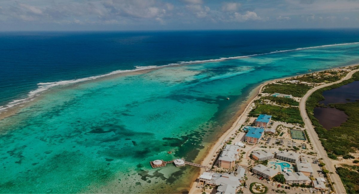 Aerial view of Grand Cayman's coastline showing turquoise reef waters, white sand beach, and beachfront properties