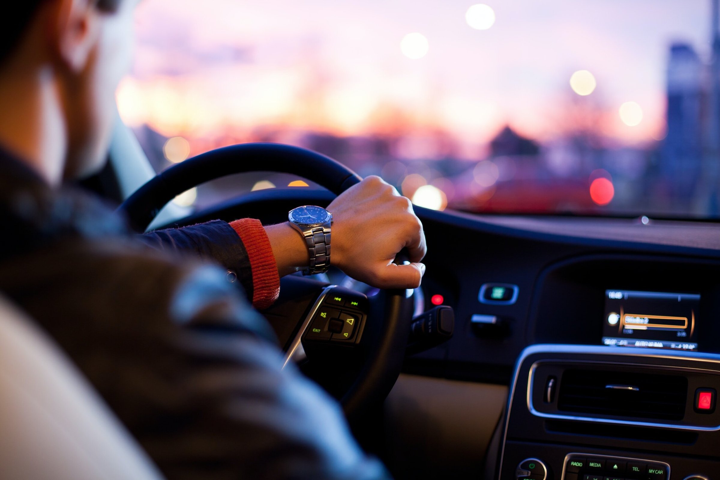 A person driving a car during sunset, with cityscape blurred in the background.
