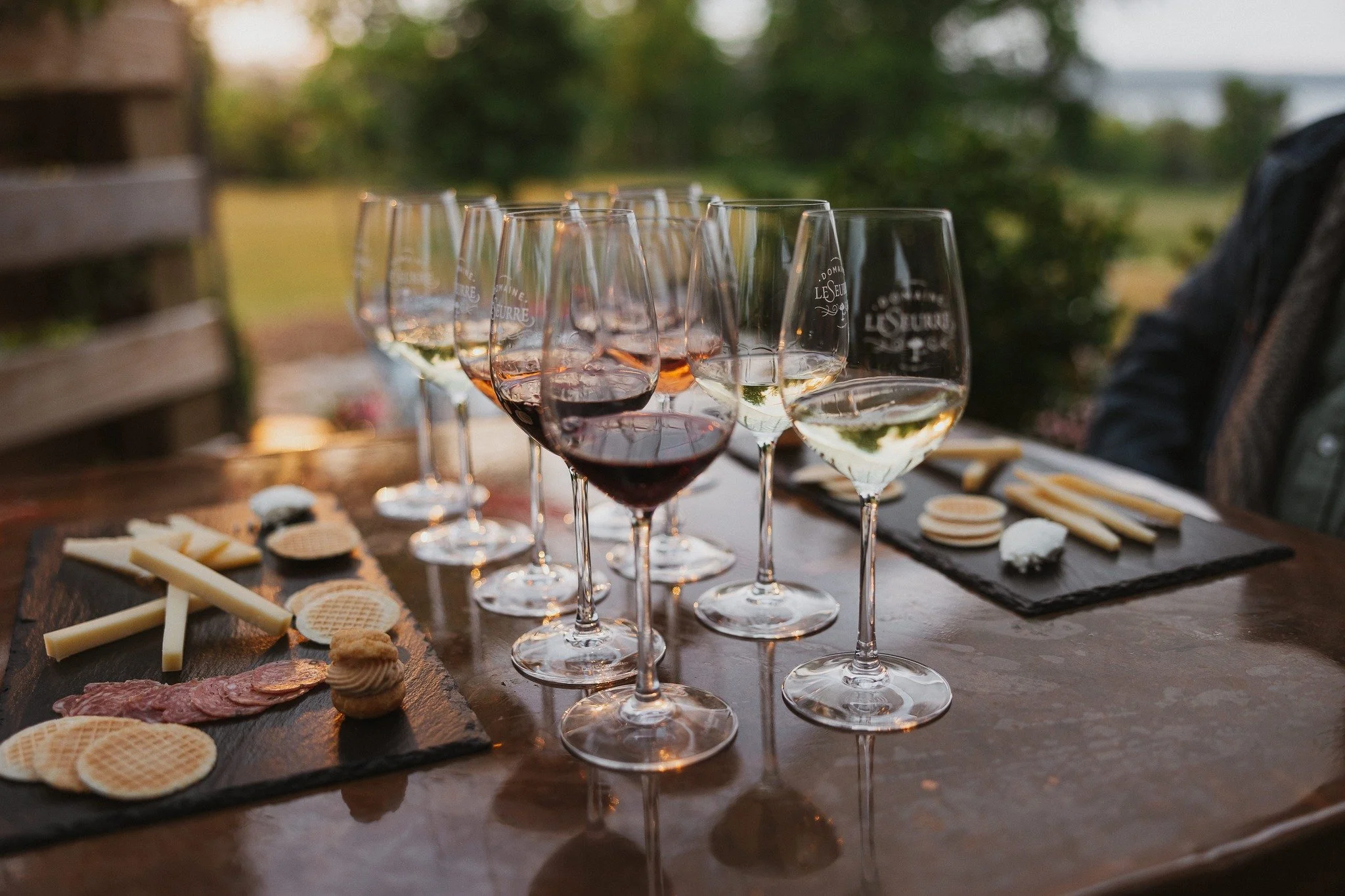 Multiple glasses of white and red wine on a wooden table outdoors, with cheese and charcuterie on black slate boards in the background.