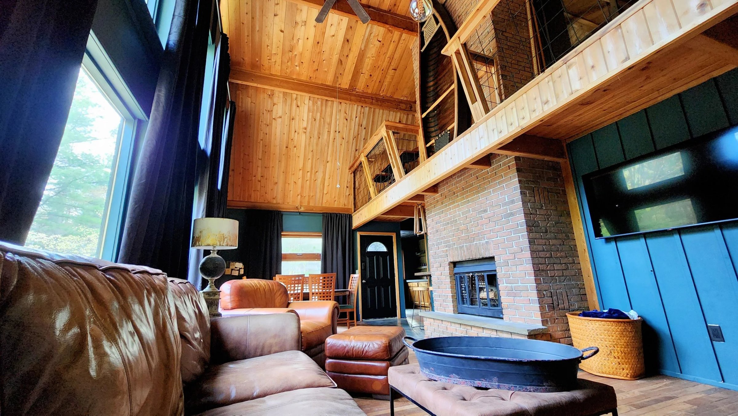 View of a cozy living room with a leather sofa, a matching armchair, a black fireplace, a flat-screen TV on a teal-paneled wall, and a rustic wooden ceiling with a loft area. Large windows with black curtains let in natural light.