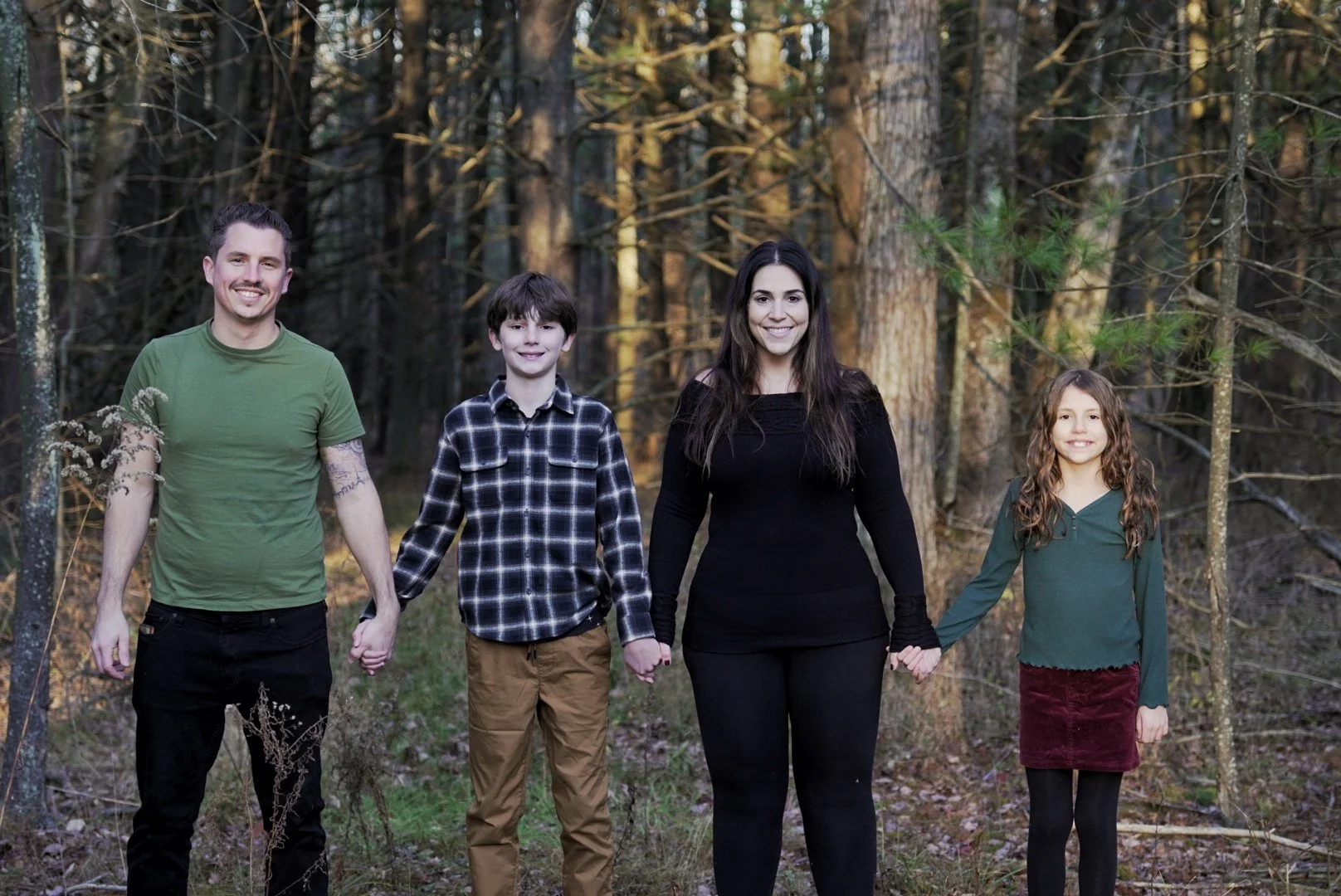 A family of five standing in a forest, holding hands and smiling at the camera. The family includes two adults, one man and one woman, and three children, two boys and one girl. The background is filled with tall trees and natural woodland scenery.