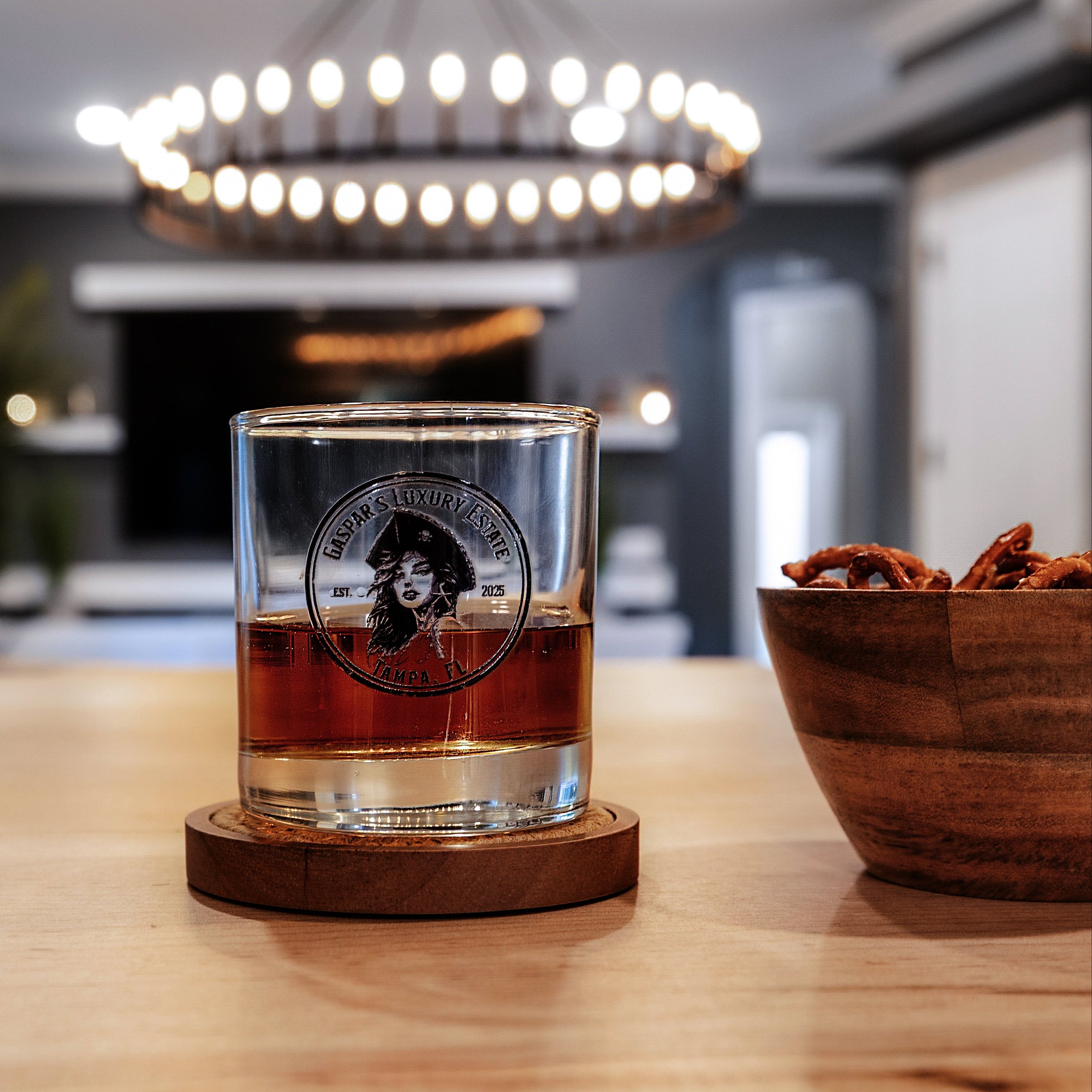 Glass of whiskey on a coaster with a bowl of snacks on a wooden table.