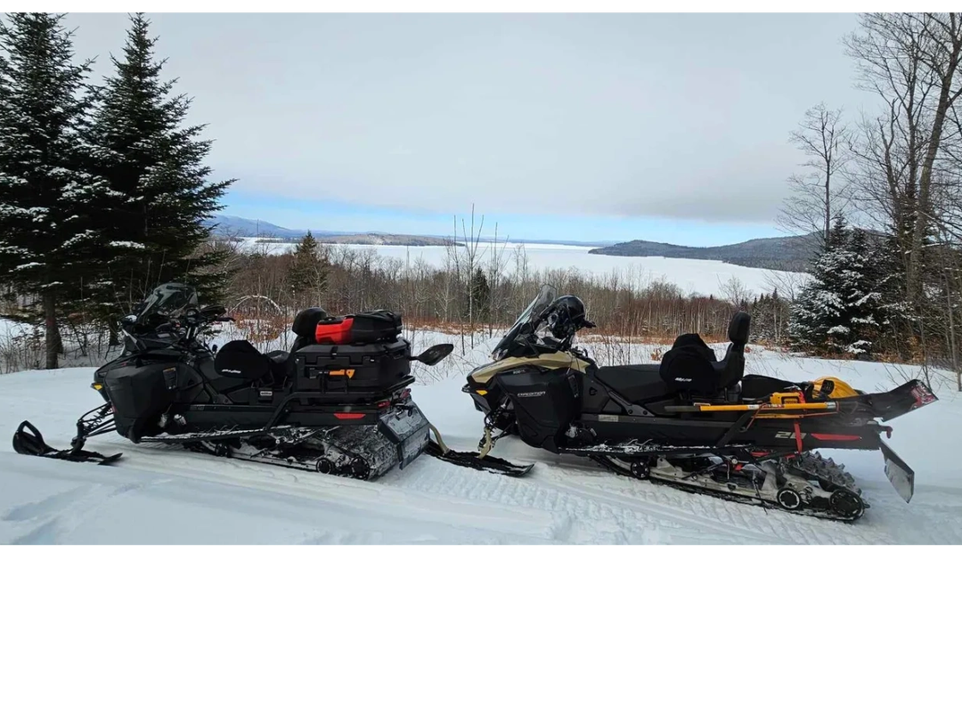 Two snowmobiles parked on a snowy trail with a scenic frozen lake view.