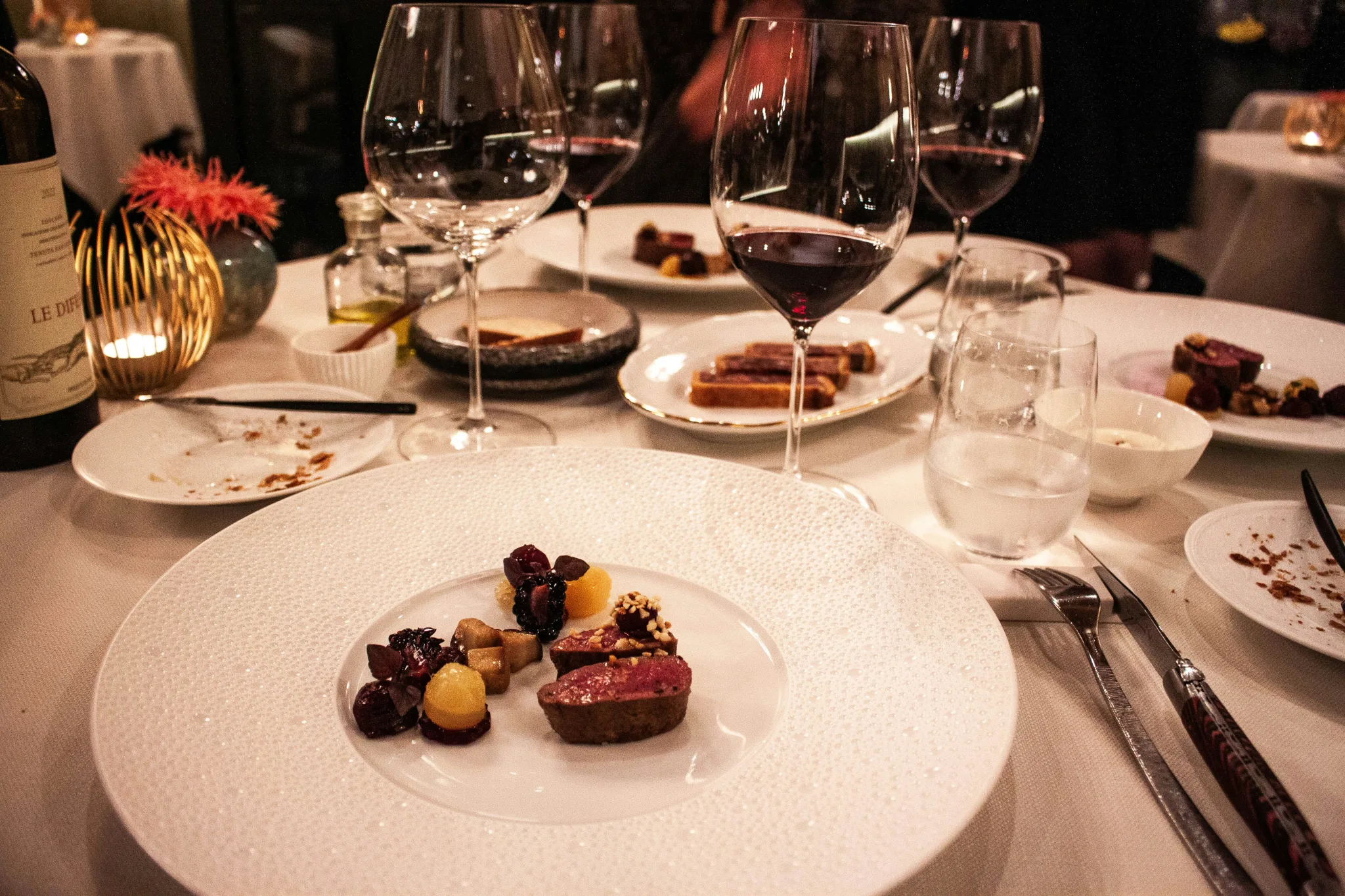 A close up photo of a table shows canapes on white plates and nearly finished glasses of red wine, with dark lighting and a candle.