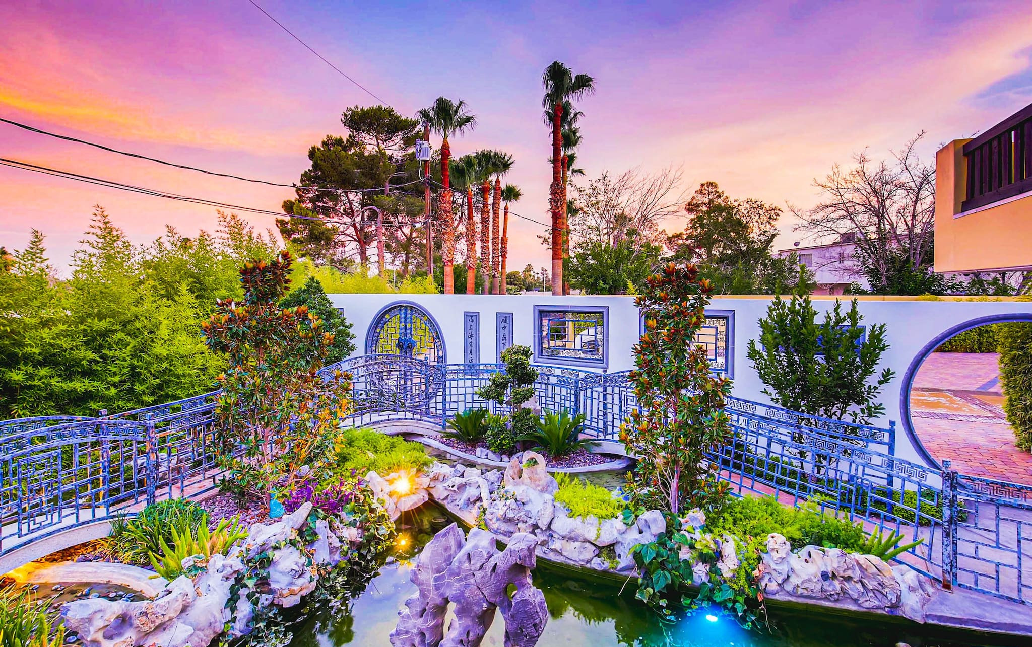 A whimsical garden is seen at a luxury Vegas villa during sunset, with a stone feature wall, bridge, pond, trees and flower gardens.