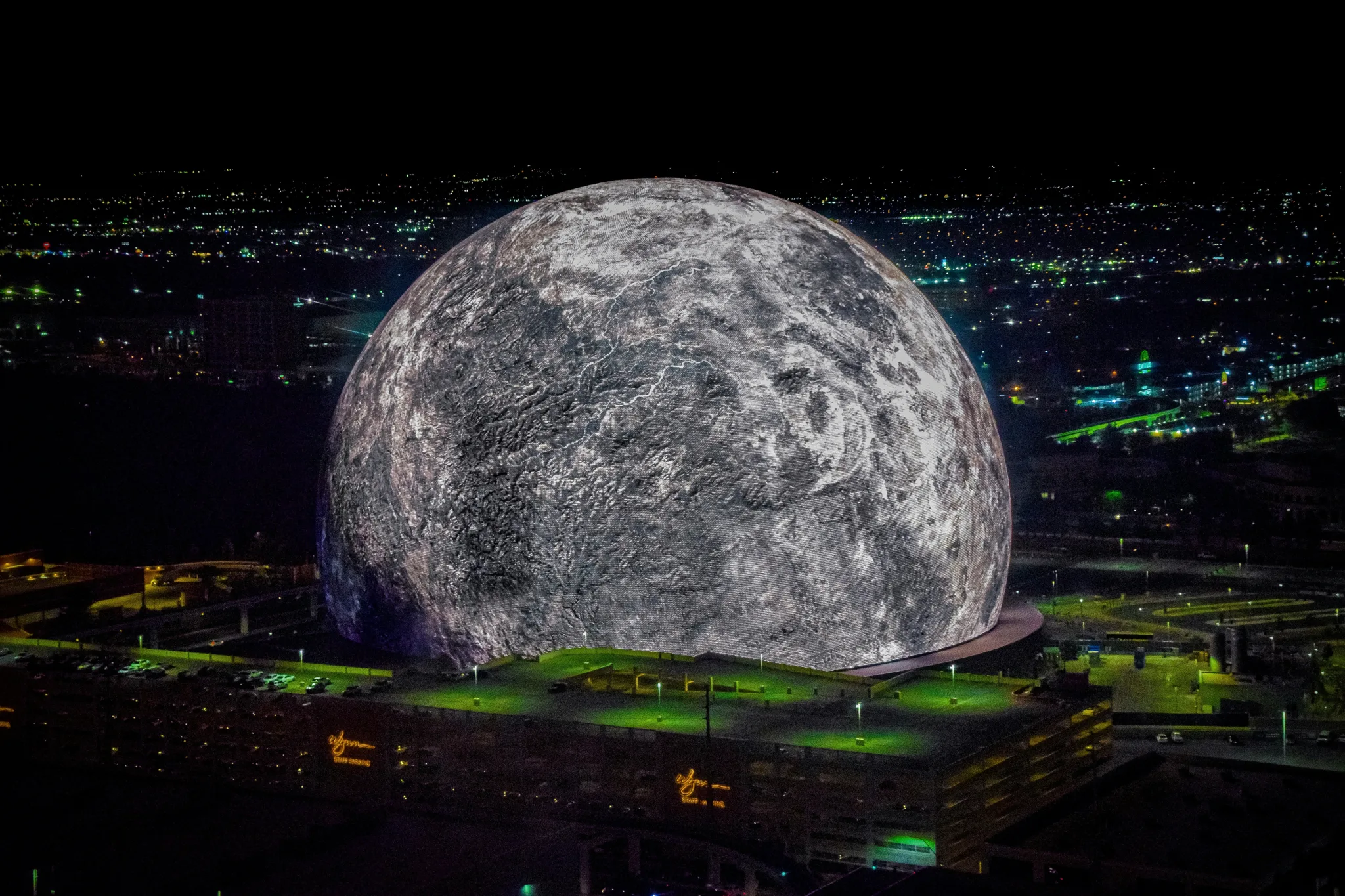 The Sphere on Sands Avenue seen at night, appearing like a giant moon with its surface textured in shades of grey, with total darkness around except for some city lights.