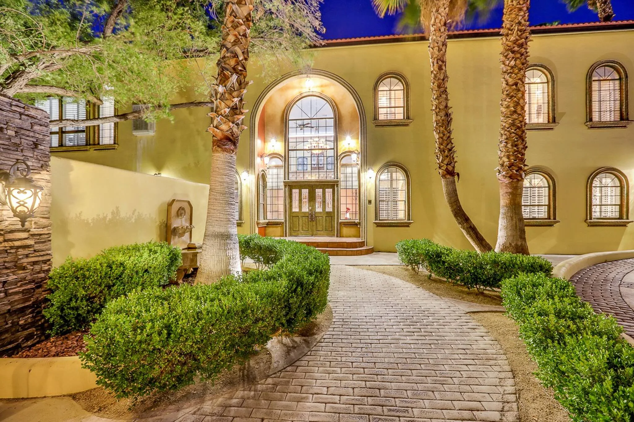 The entry way to D&D Mansion in Vegas is grand and inviting, with a brick pathway lined with green shrubs and palm trees, and a huge archway door surrounded by yellow stucco