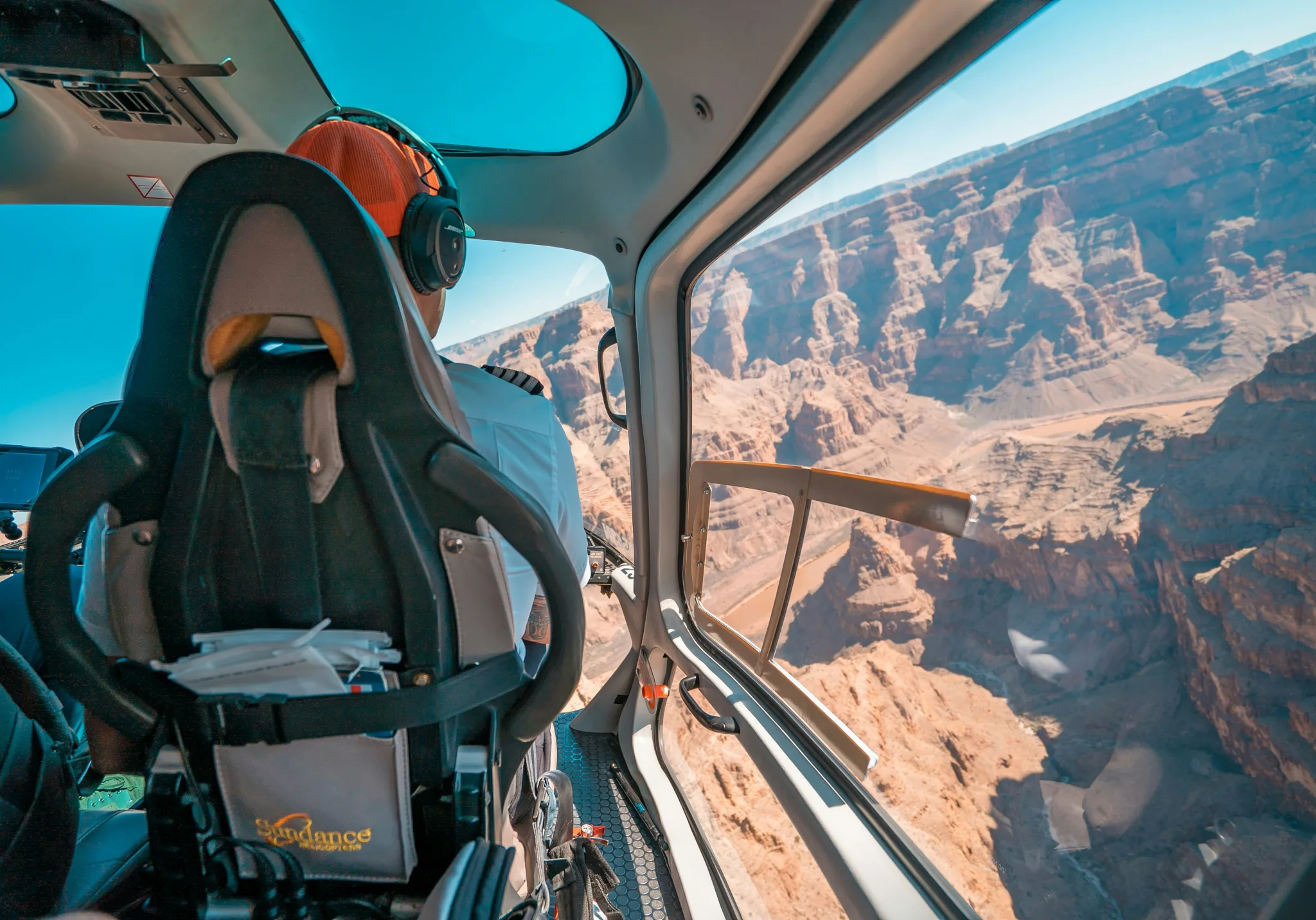 From the inside of a helicopter over the Grand Canyon, we can see the seat ahead with the pilot, and a full glass side offering panoramic view of the brown rock canyon.
