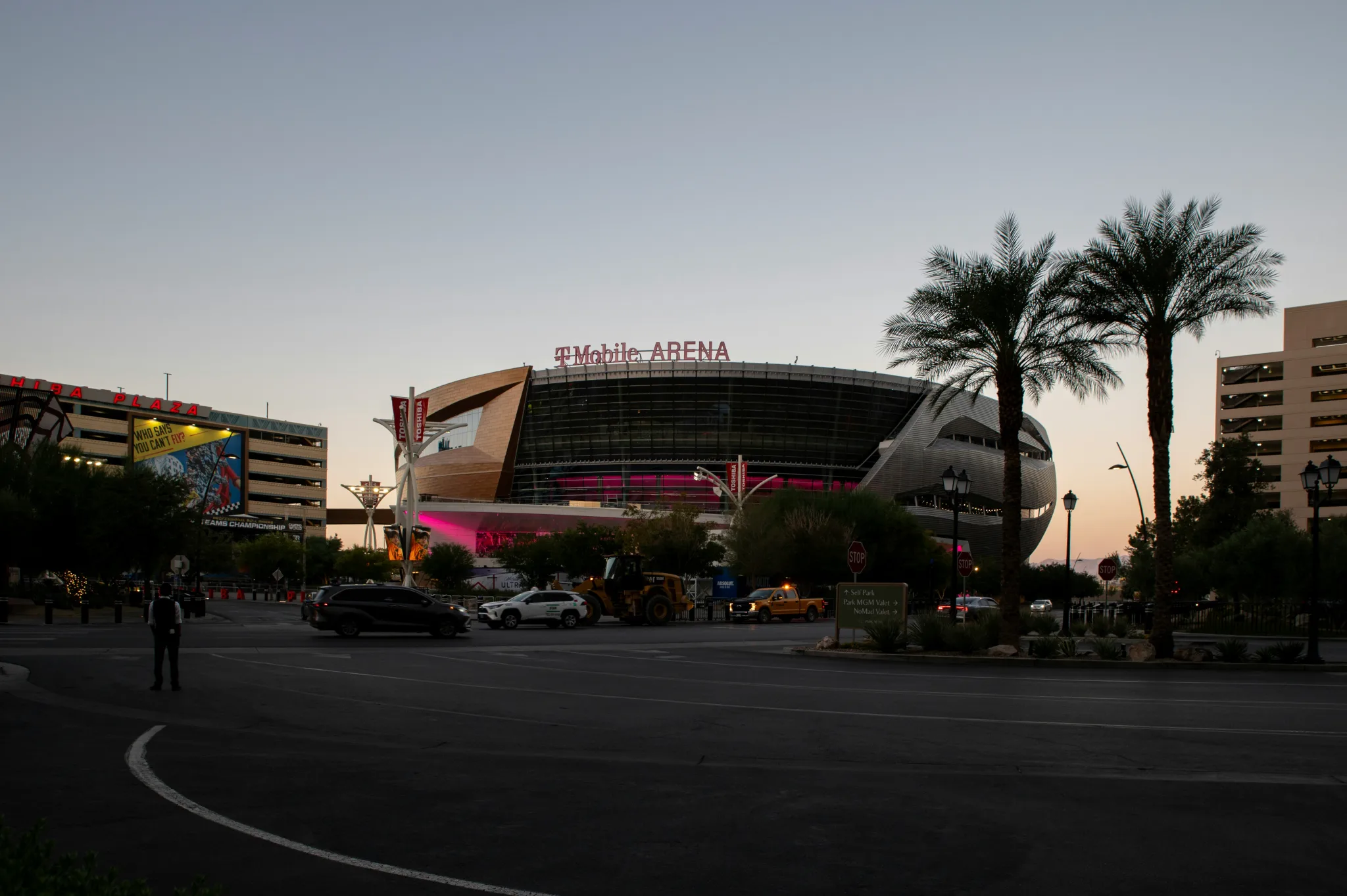 A round, flat-topped arena  is becoming a shadow as the sun goes down. The sign reads "T-Mobile Arena" nad some cars and palm trees can be seen on the Vegas road in front of it.