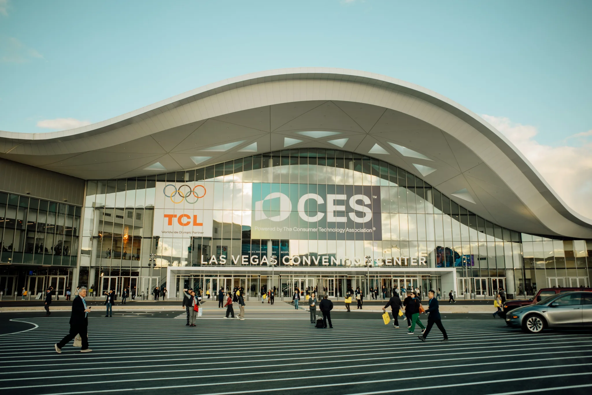 The Las Vegas Convention Center, a modern grey and glass building with curved rooftop, is seen with people walking by, with a giant CES banner on the front of it