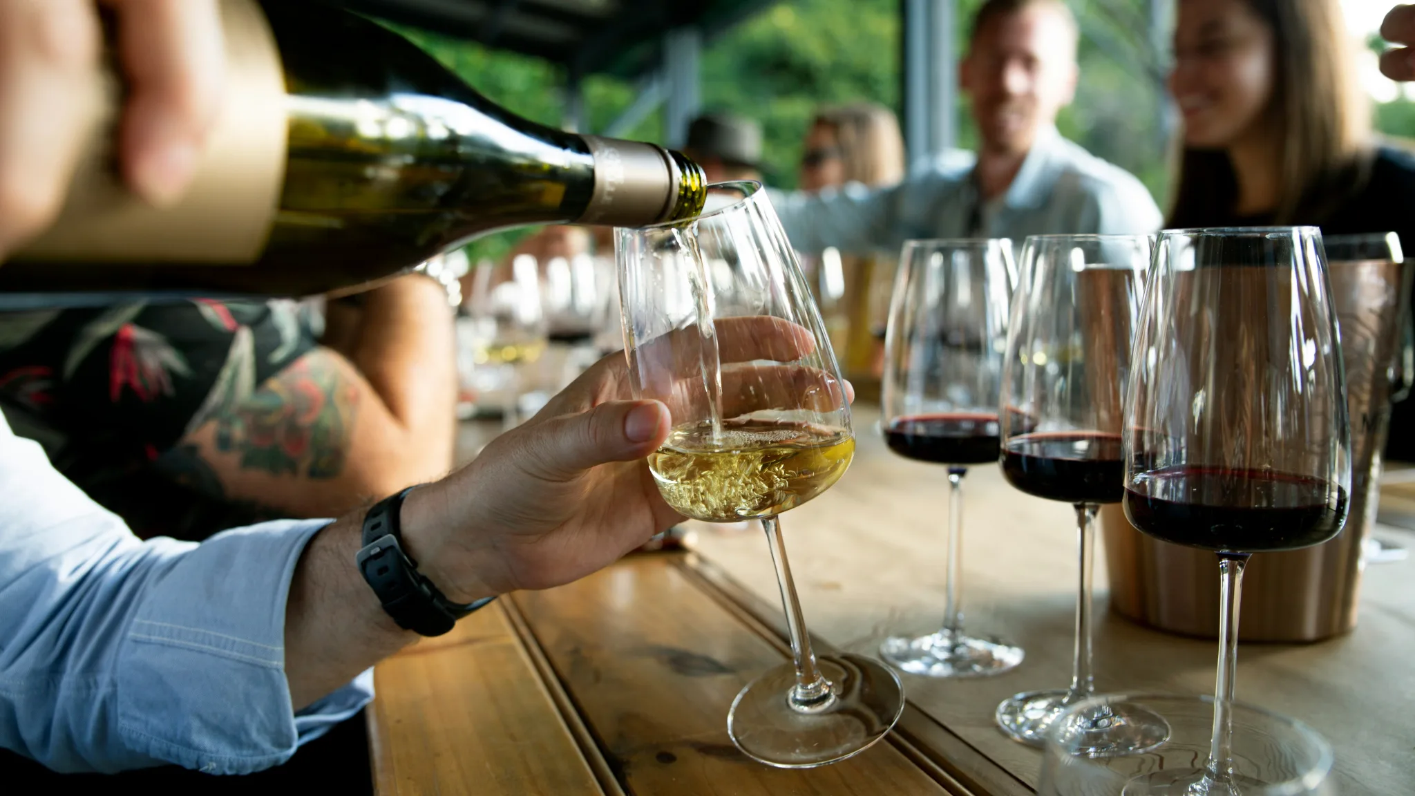 POV from the sommelier who is pouring a white wine into a wine glass, with 3 glasses of red ahead of it, and faces of two couples waiting at the bar.