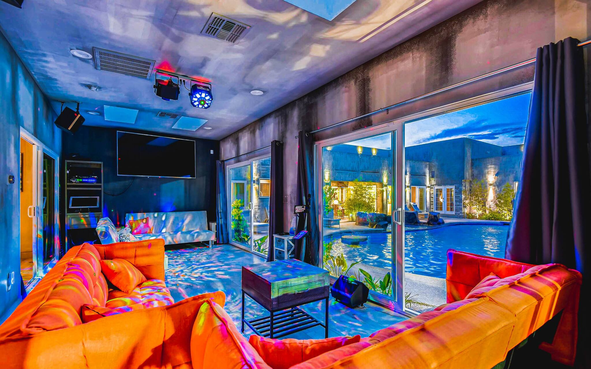 A living room in R&R Mansion features bright orange couches, a flat screen TV and party lights, and looks out at the villa's pool.