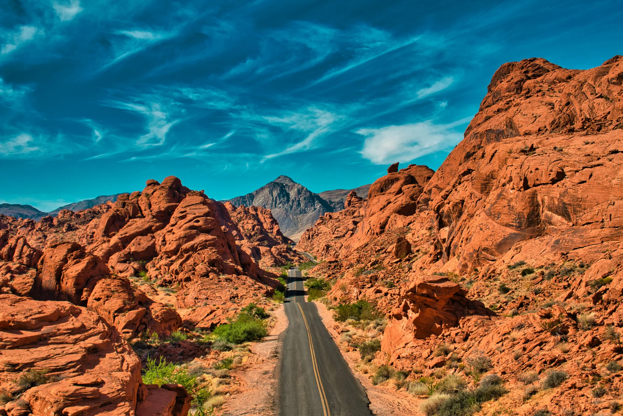A black pavement road rolls up and down through a valley of red rock formations with a dramatic dark blue sky and mountains in the distance.