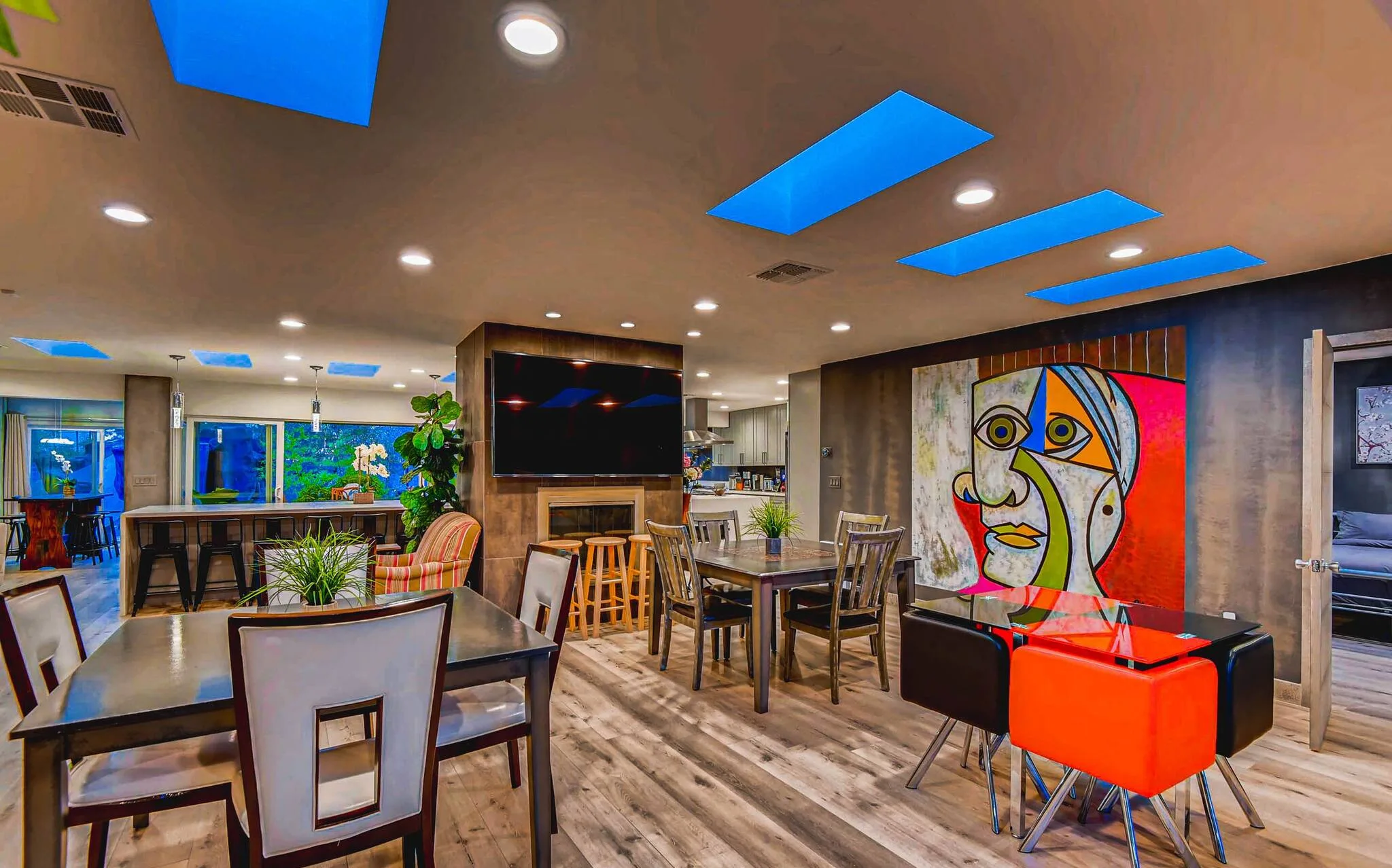 Funky mismatched furniture creates a fun, colorful and modern design in the living area at R&R Mansion in Vegas.  Three tables with chairs of different shapes and colors are in the foreground, a large red, white and yellow modern art photo of a jigsaw-like face stands out on the wall, and in the background, a large screen TV and kitchen island can be seen.