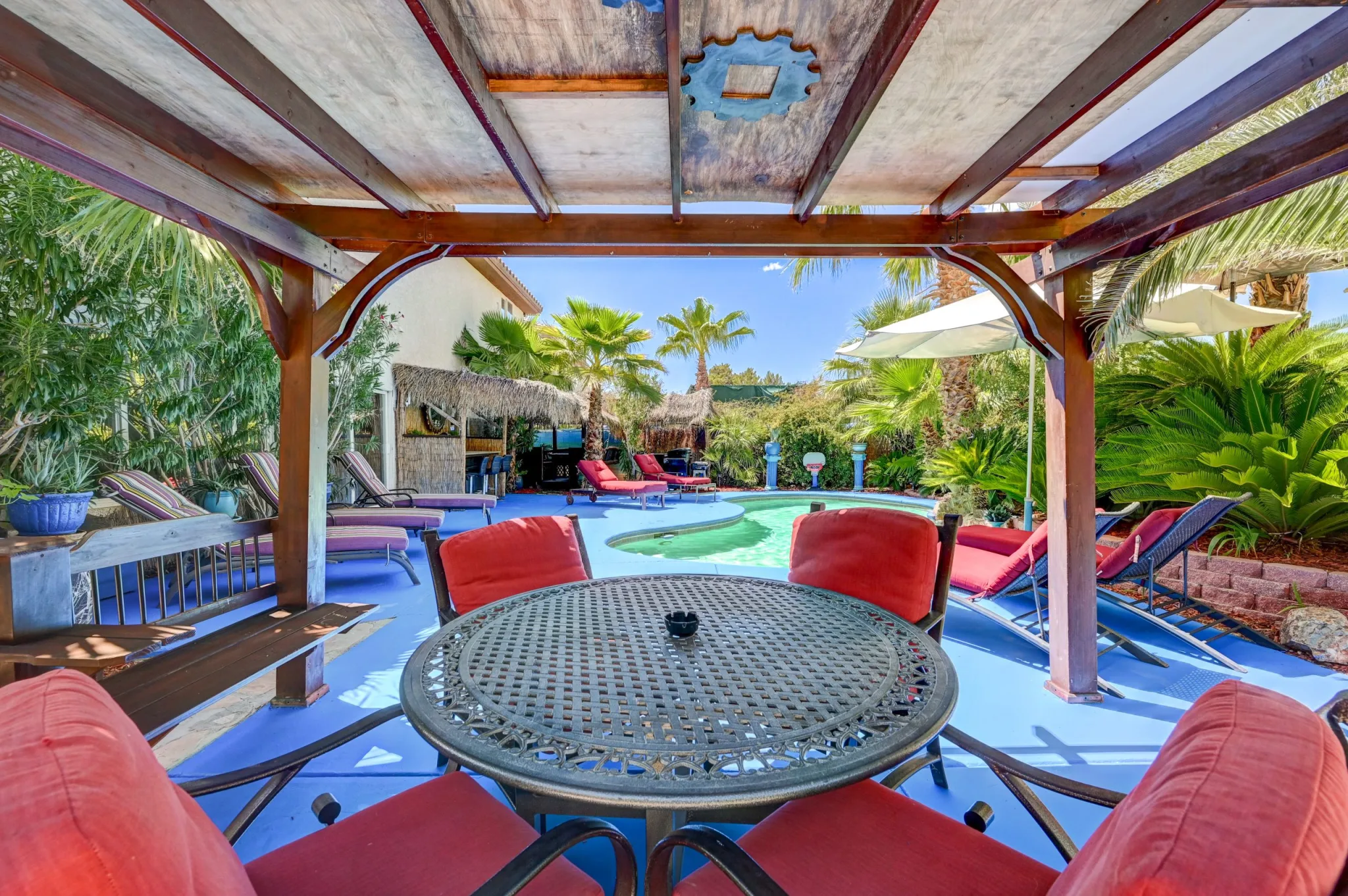 From a shaded area with outdoor table and chairs, a resort-style pool, more seating, and greenery are seen in the backyard of Paradise Estate, a rental mansion in Vegas.
