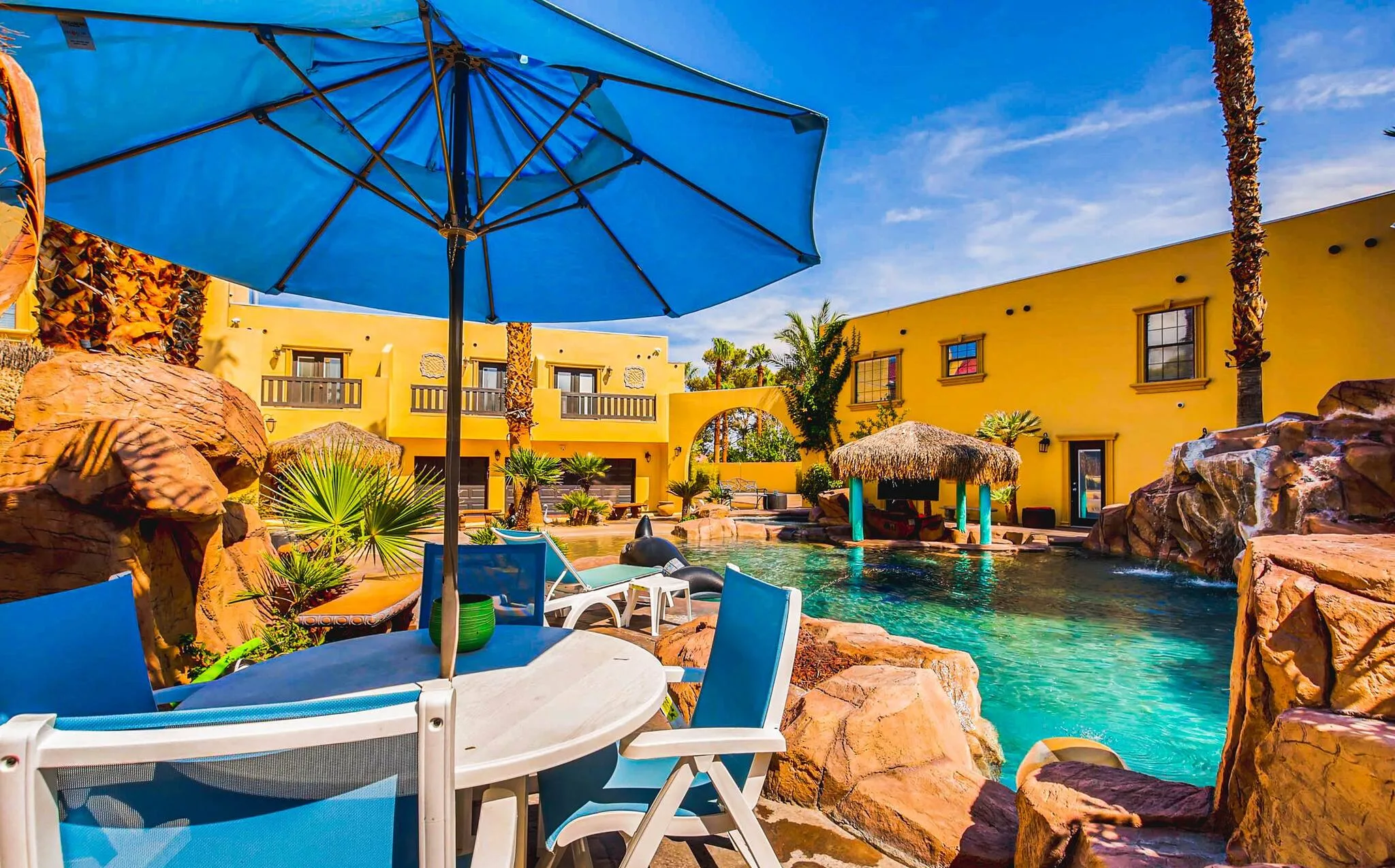 An outdoor table with umbrella and chairs are in the foreground, ahead of stone features surrounding a tropical style pool at D&D Mansion in Vegas. The large stucco house can be seen behind the aquamarine pool.
