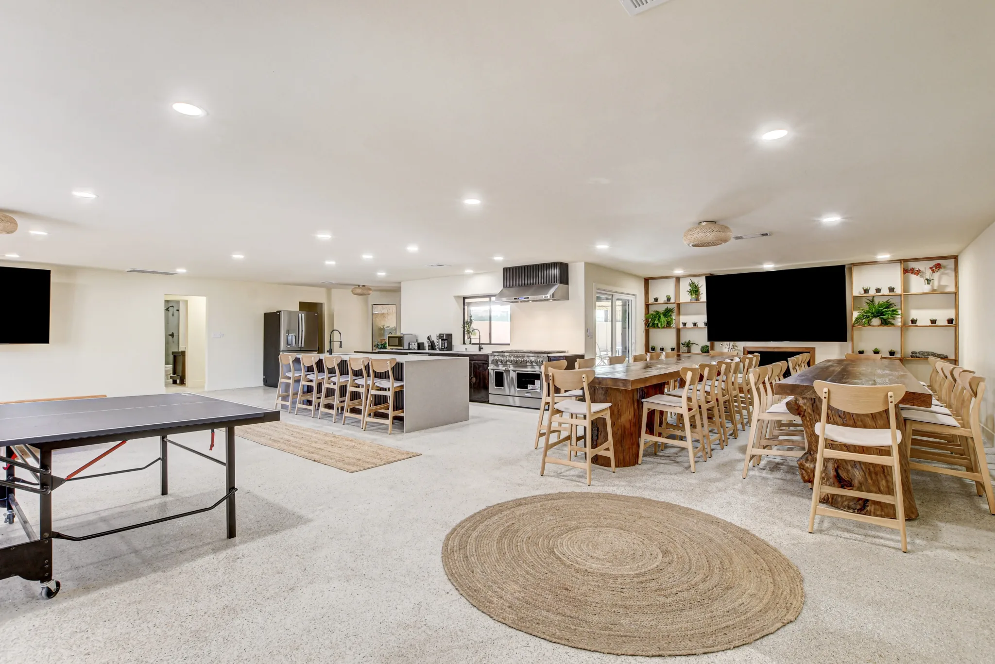 A wide angle shot of the open living space in Zen Paradise, a villa in Vegas, featuring dozens of seats at large dining tables, a kitchen island with barstools, a pristine modern kitchen, flatscreen TV, and pingpong table.
