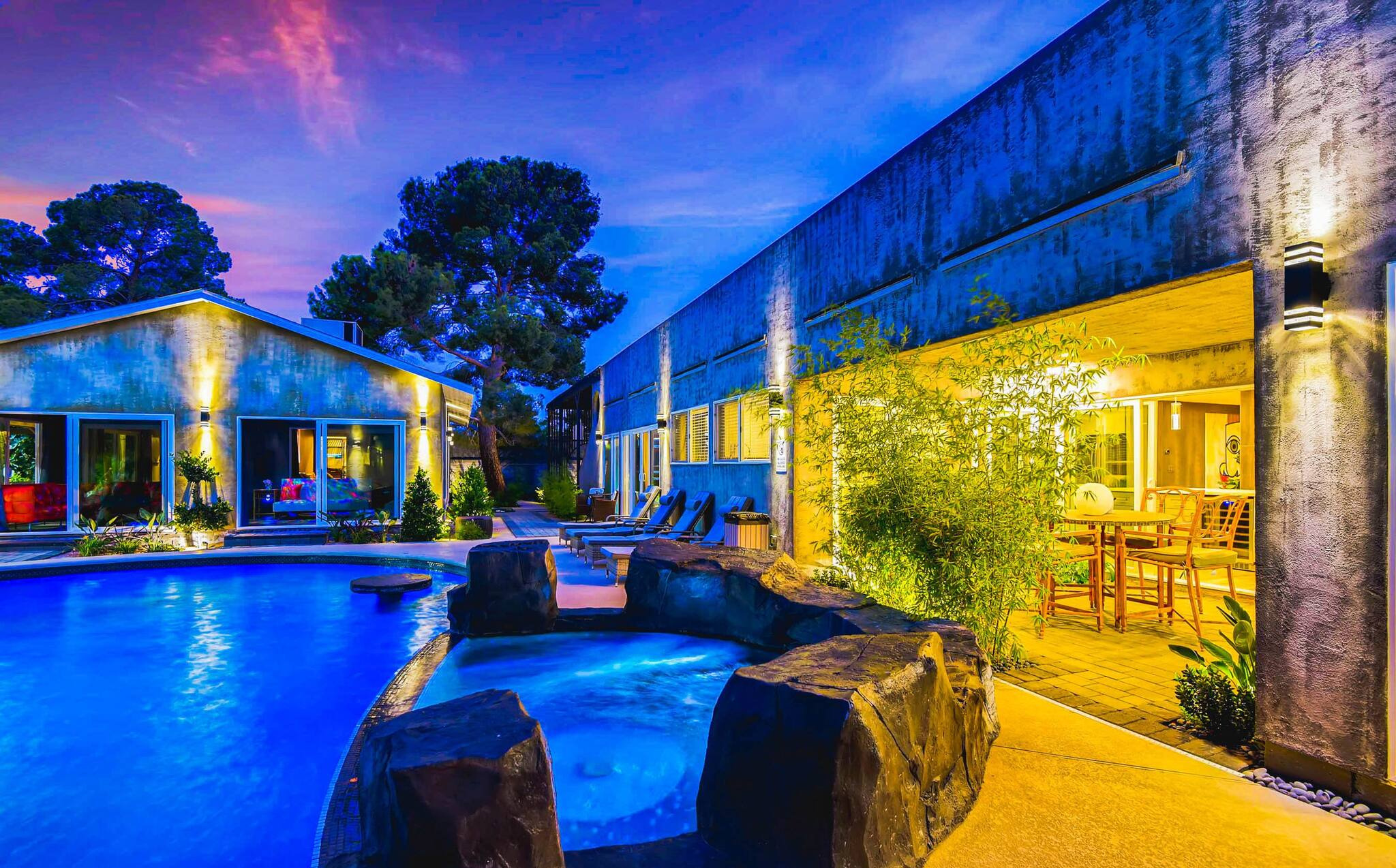 Hues of blue reflect off a pool at Paradise Estate onto the villa and warm yellow light emanates from the covered outdoor lounge, showing a cozy and luxurious rental space in Vegas.