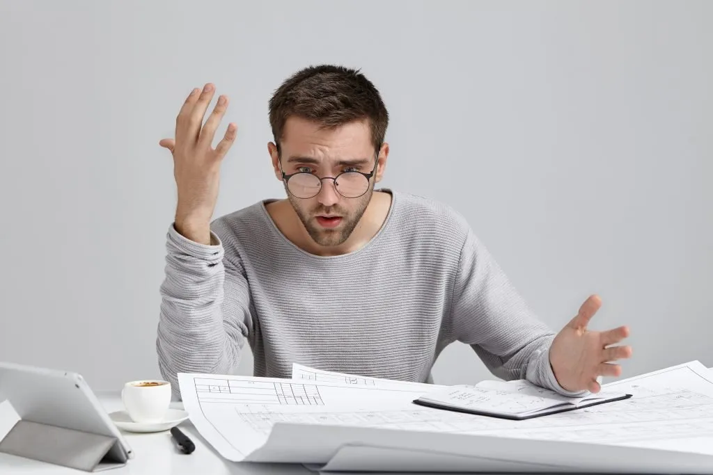 Puzzled and frustrated young architect in casual sweatshirt and trendy glasses feeling indignant, looking at drawings on his desk, realizing how much work he has to do. Human facial expressions