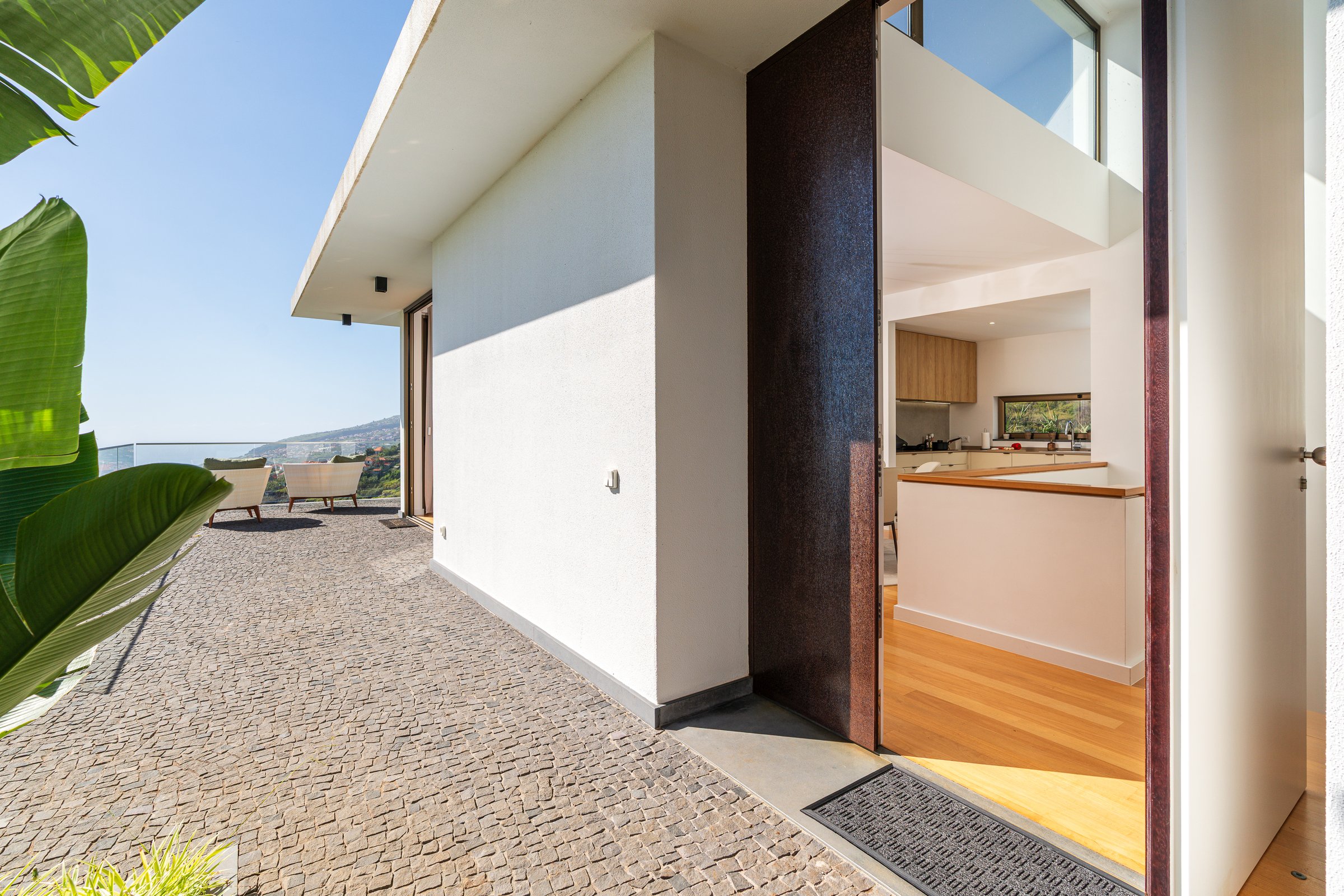 Entrance of a contemporary villa in Madeira with an open door, bright interior, stone terrace, and wide views over the sea and hillside.