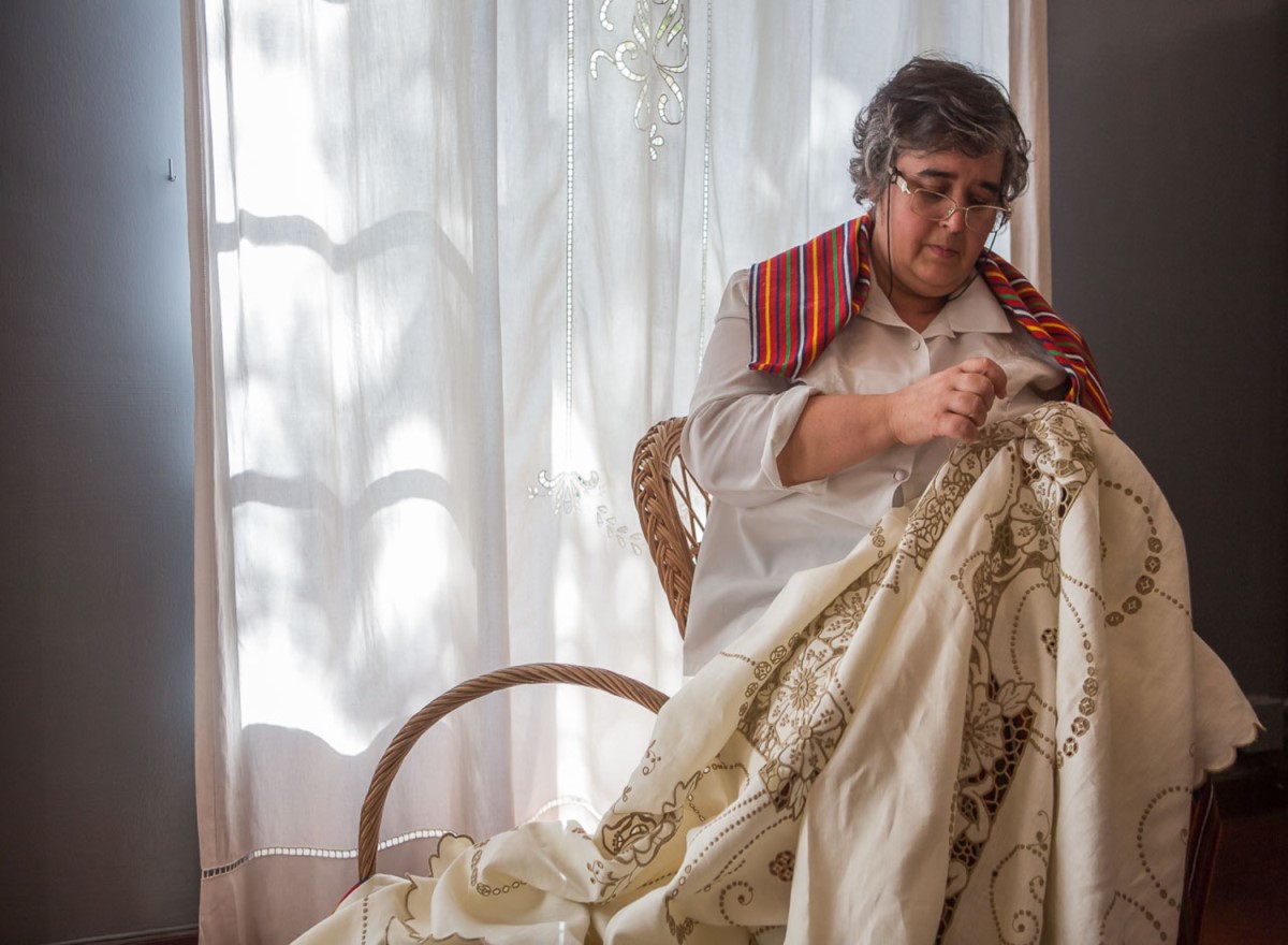 A woman seated by a sunlit window, wearing glasses and a striped shawl, hand-embroidering an ornate linen cloth, reflecting the enduring craft of Madeira embroidery.