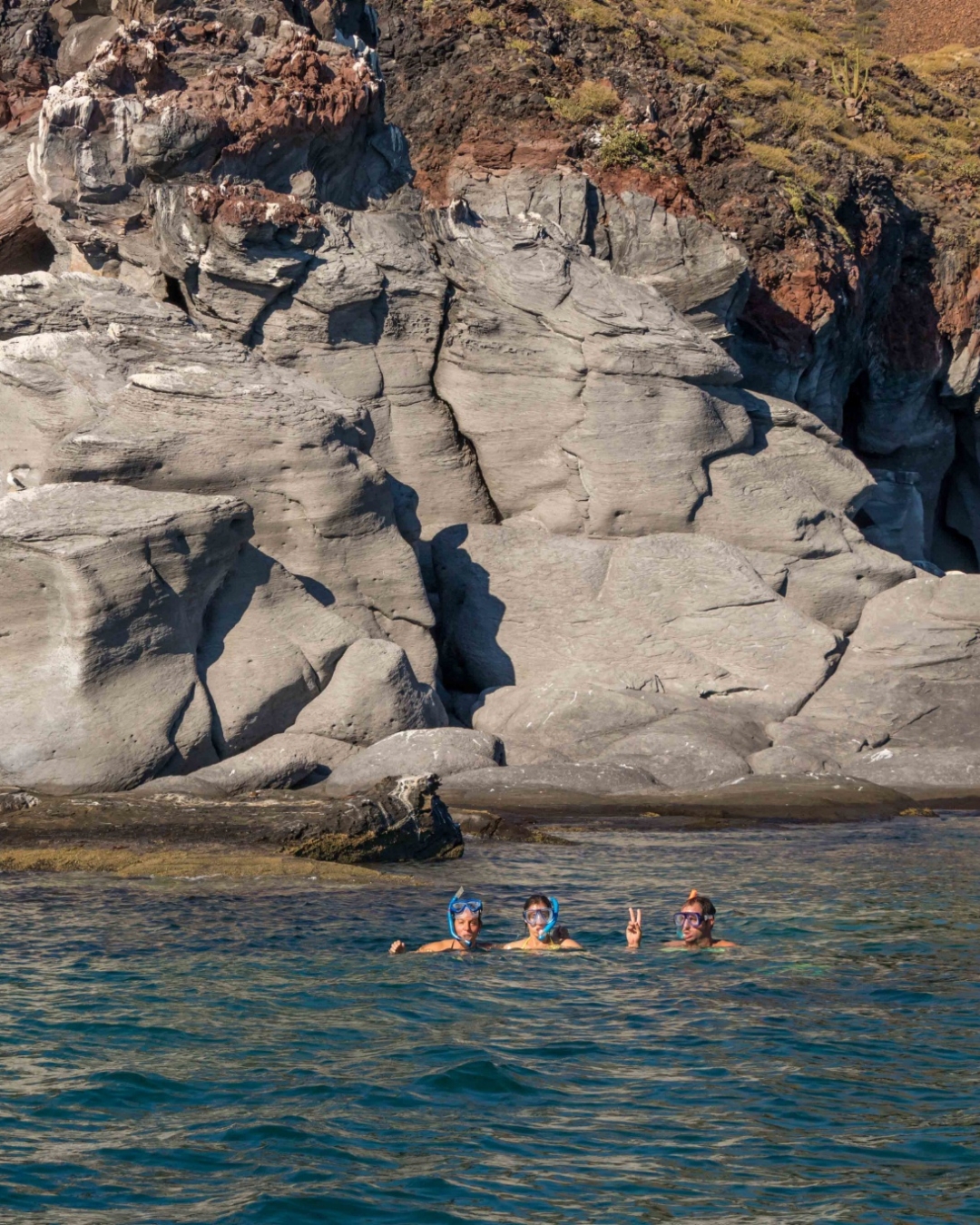 Three snorkelers in masks floating in front of volcanic rock cliffs in the Sea of Cortez