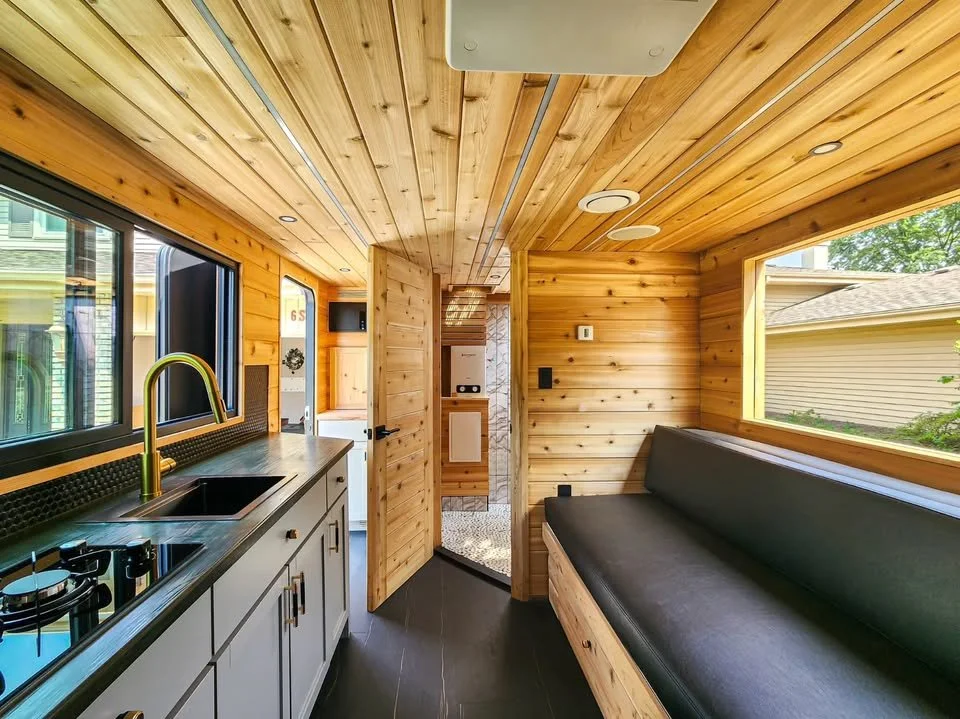 Interior of a tiny wooden house with a kitchen on the left, black countertop, sink, stove, and large window. On the right, a built-in black cushioned bench by another window. Wooden walls and ceiling, door leading to another small room with a washer