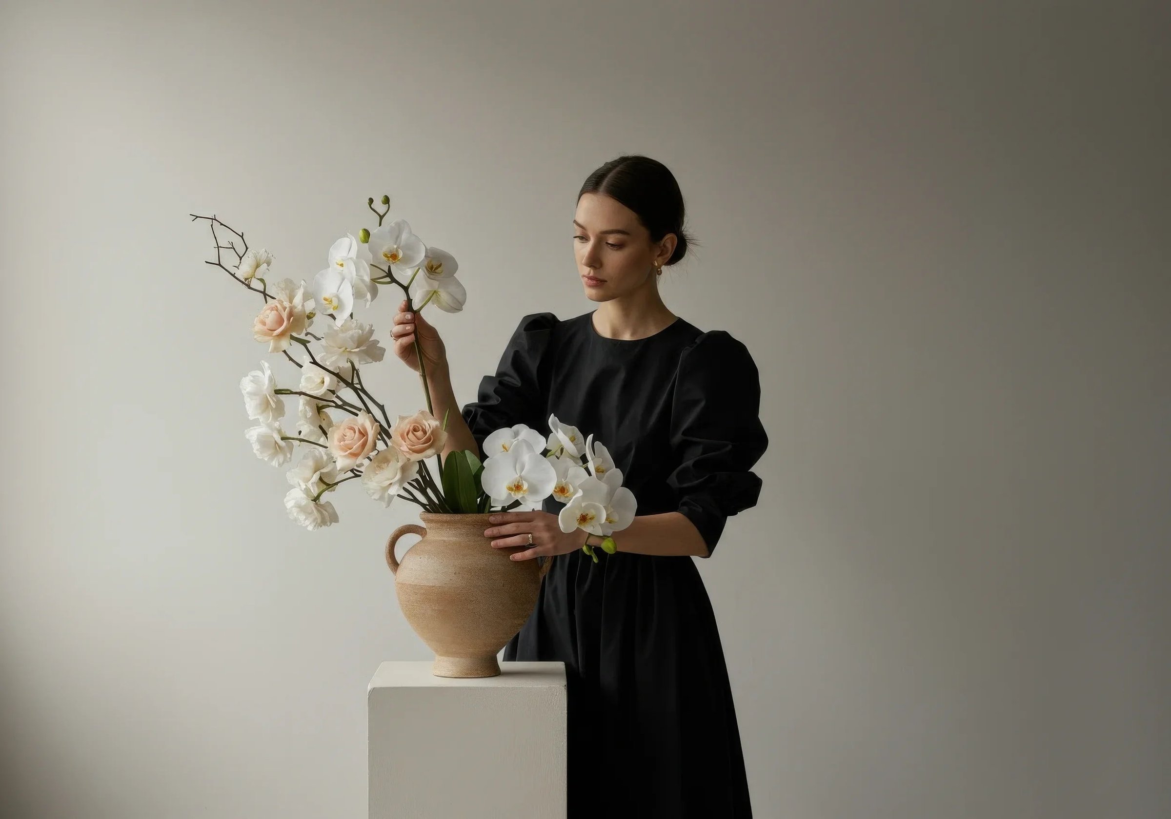 A woman in a black dress arranging a large bouquet of white orchids and pale pink roses in a beige ceramic vase on a white pedestal.