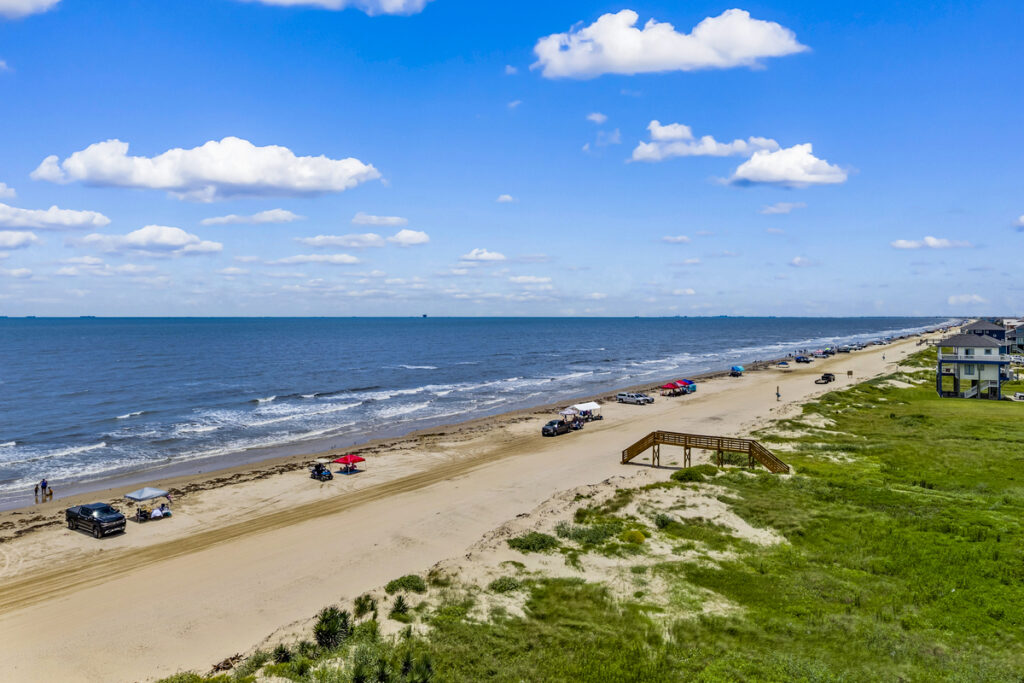 Aerial view of Bolivar Peninsula Texas showing trucks parked on the sand, beach access boardwalk, coastal dunes, and vacation homes along the shoreline