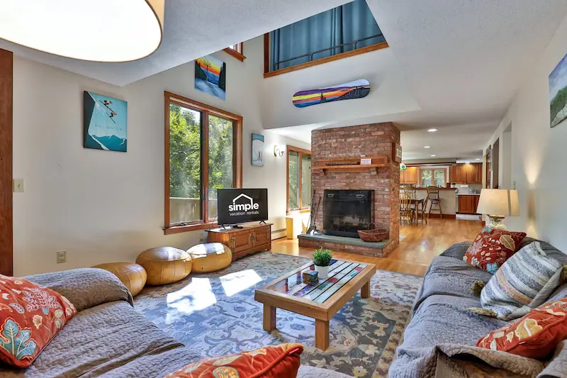 Living room of a vacation home near Killington, VT, with sofas surrounding a fireplace and a smart TV. Large windows show forest views.