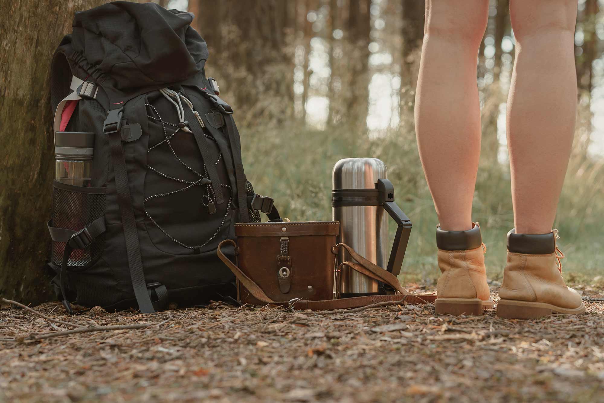 Hiker with boots stands beside a backpack, thermos, and leather bag on a trail to Greeter Falls.
