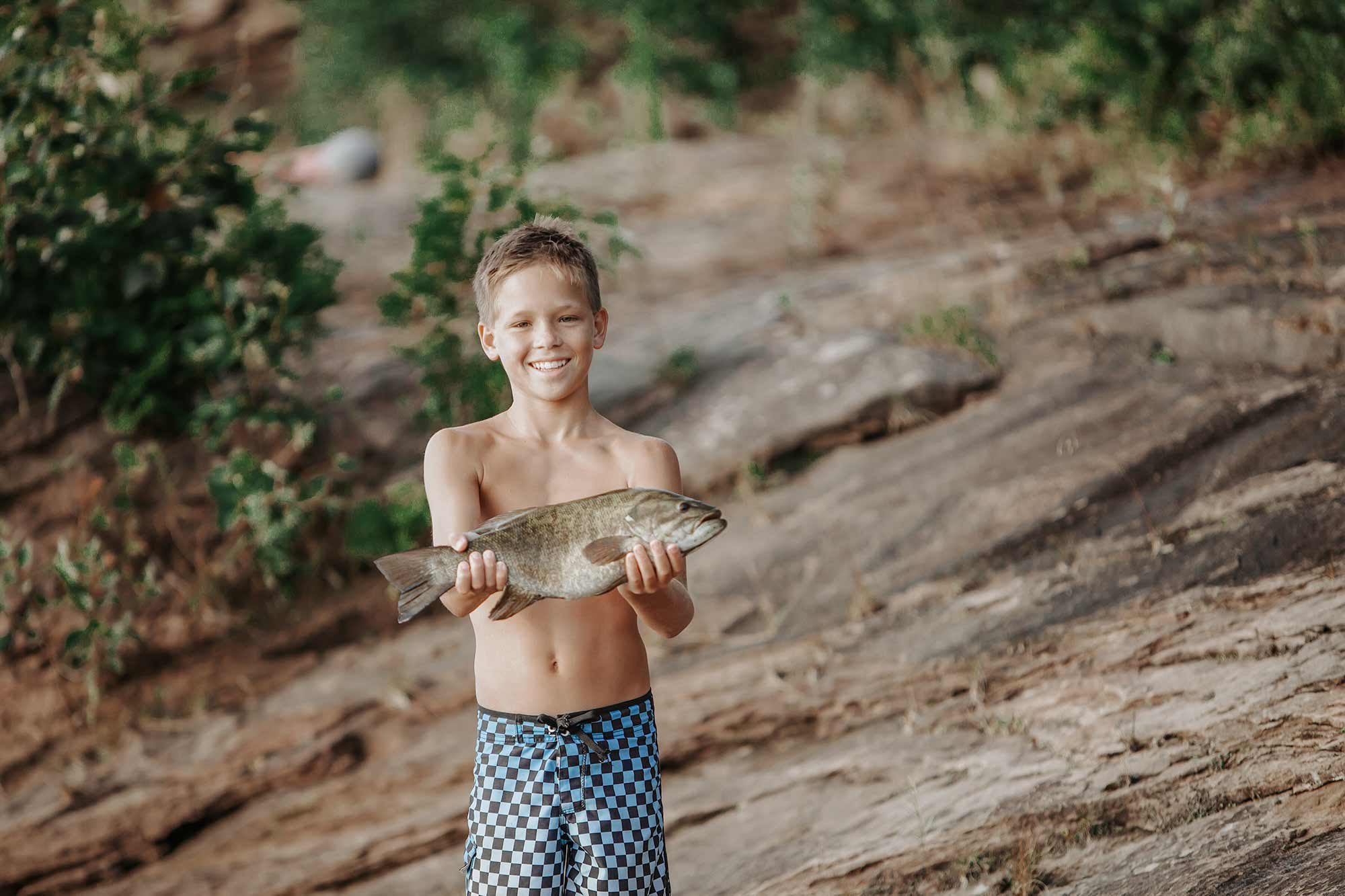 Smiling boy proudly holding a freshly caught smallmouth bass on the rocky shoreline of Center Hill Lake, perfect for beginner anglers.