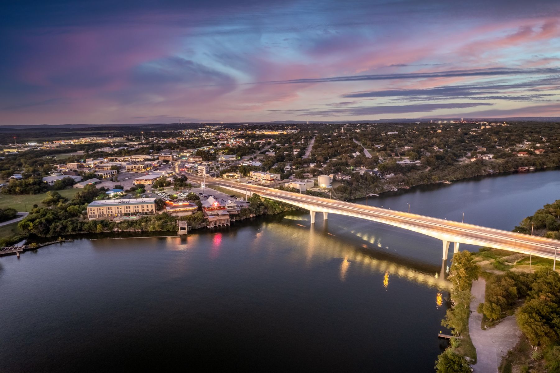 A beautiful aerial twilight view of the bridge crossing Lake Marble Falls in the Texas Hill Country. The glowing city lights and vibrant purple sunset reflect on the water, highlighting the scenic landscape and charm near Casago Highland Lakes vacation rentals.