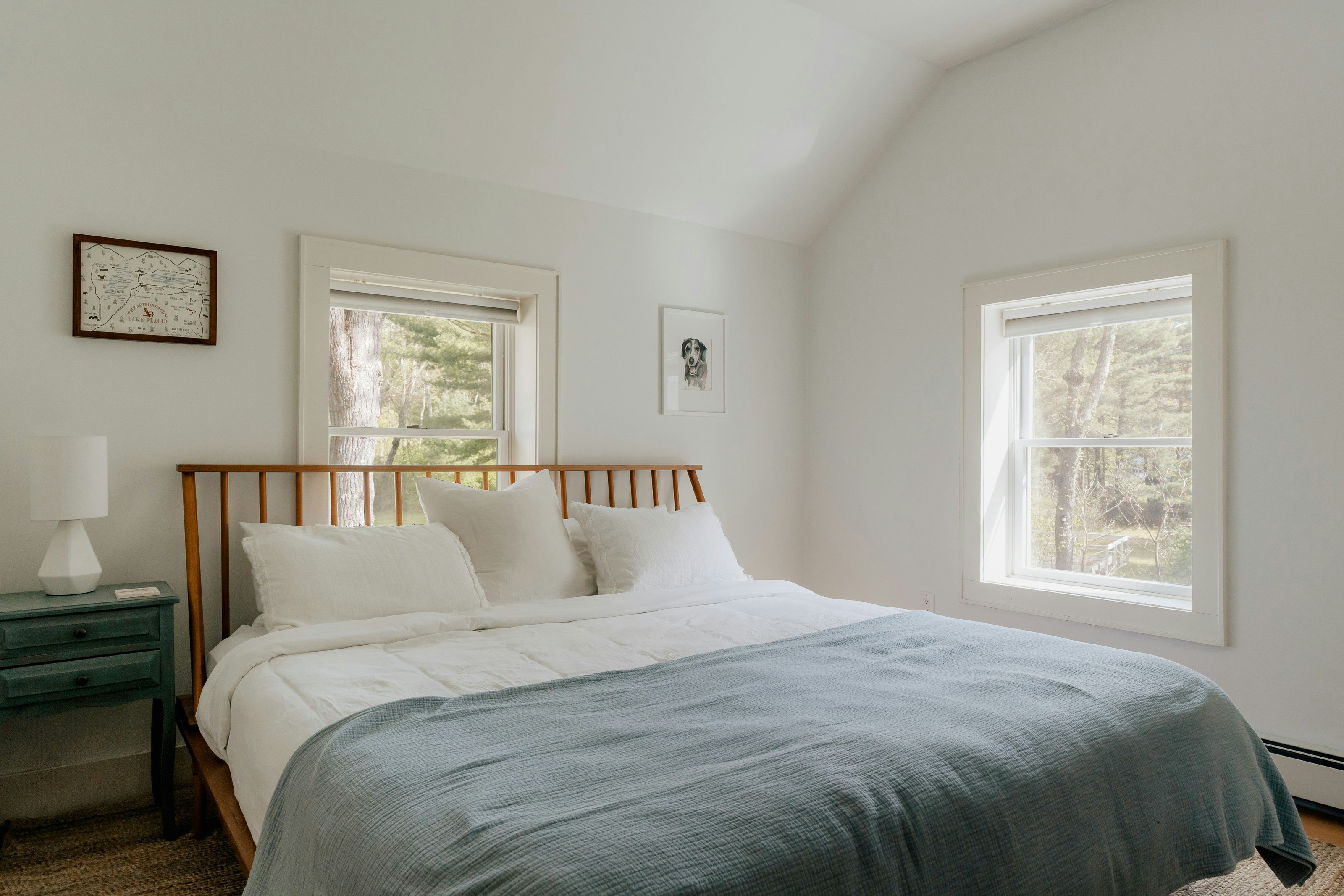 A bright, airy vacation rental bedroom with vaulted ceiling, sage-green duvet and forest views through the windows.