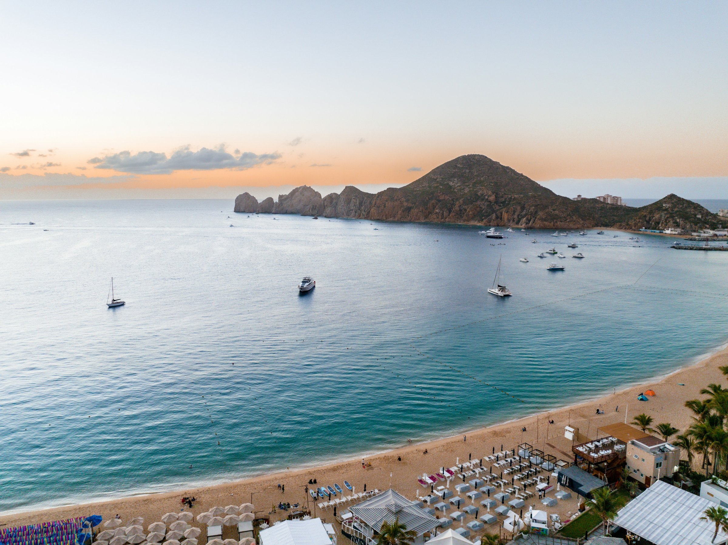 Medano Beach in Cabo San Lucas at dusk during the December holiday season, beach clubs lit along the sand with Land's End rocks in the distance