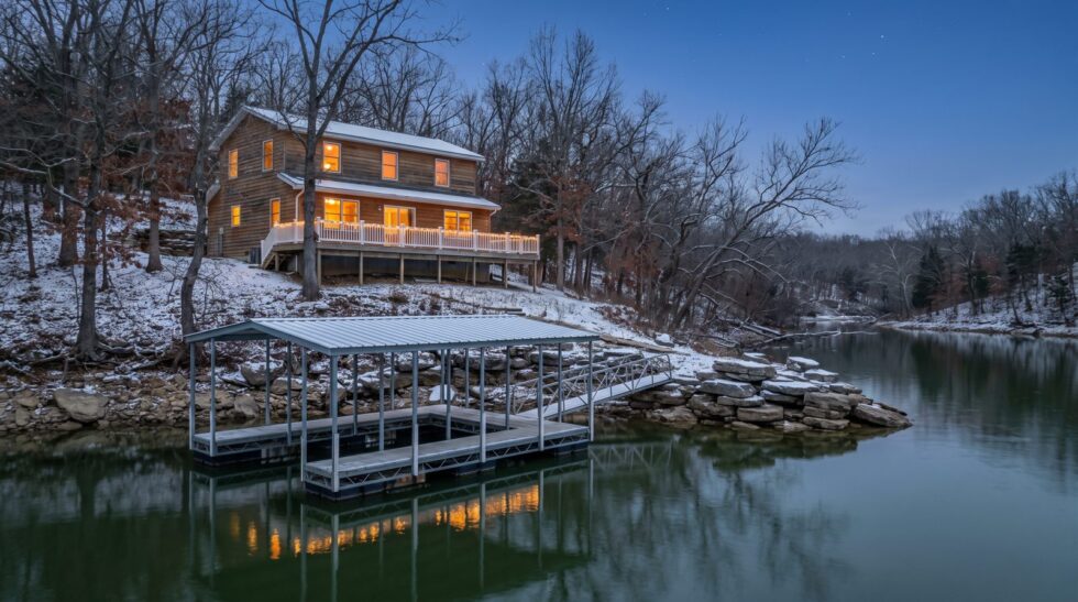 Lake house with glowing windows and holiday lights above a snowy shore and steel dock at Lake of the Ozarks
