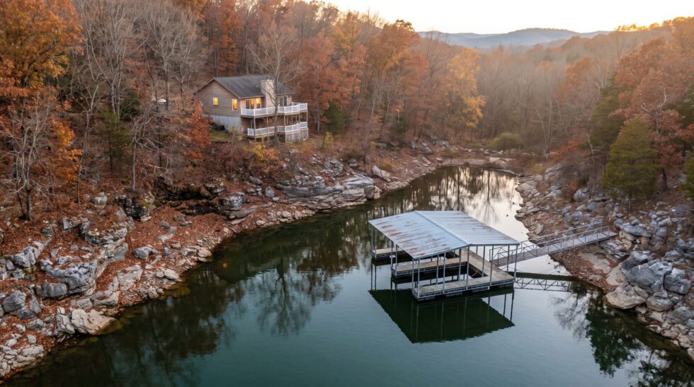 Quiet late fall cove at Lake of the Ozarks with a lake house, empty steel dock, and rust-colored trees