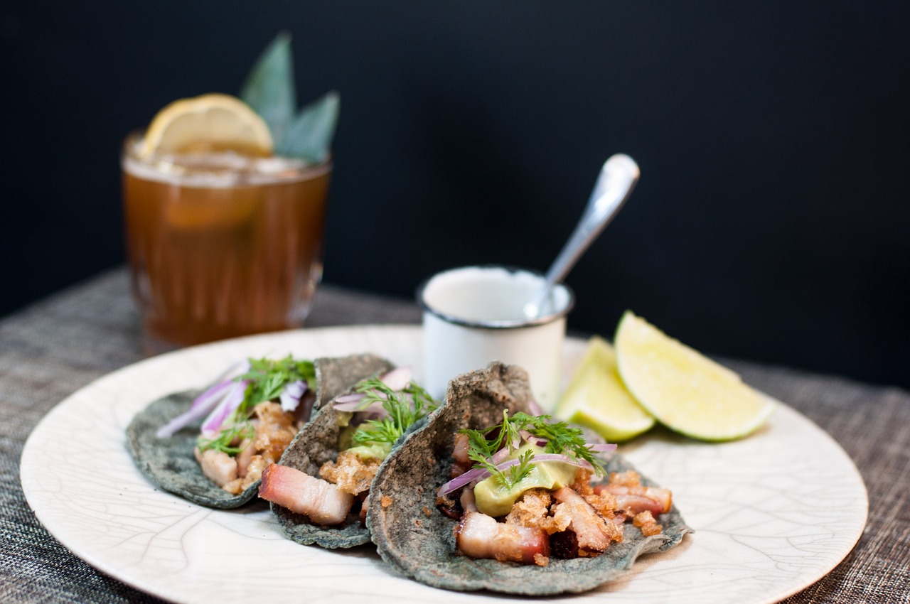 Tacos de maíz azul con cerdo, cebolla morada y hierbas en un plato blanco, con gajos de lima y un cóctel
