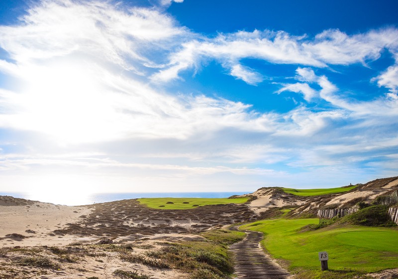 Calle de un campo de golf entre dunas de arena y el océano Pacífico bajo un amplio cielo azul