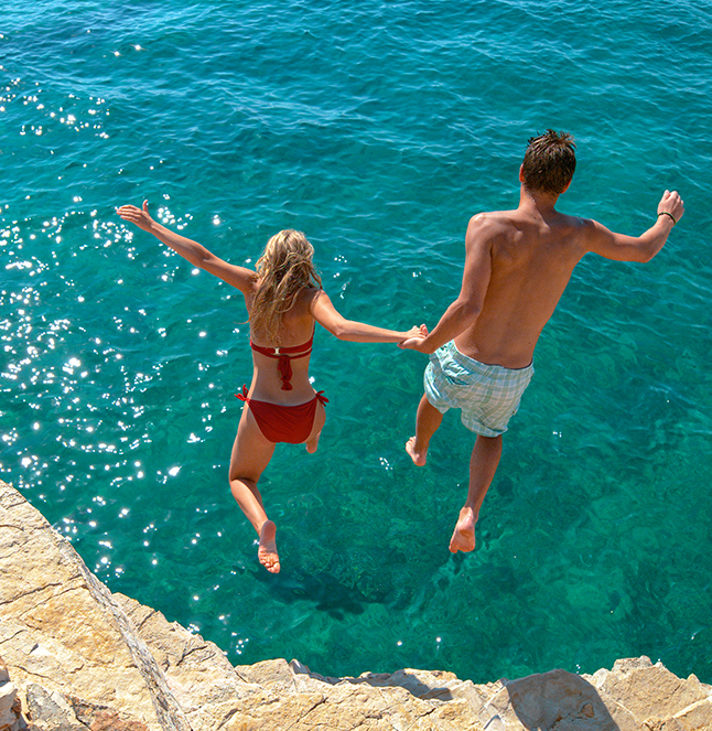 Pareja que salta de la mano desde un saliente rocoso hacia las aguas cristalinas de color turquesa