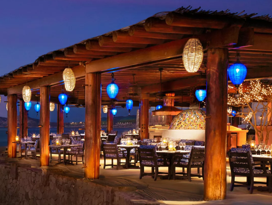 Restaurante al aire libre frente al mar al atardecer, con faroles azules colgados del techo de una palapa y mesas preparadas con vistas al mar
