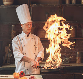 Chef in white uniform flambéing a pan over an open flame in a restaurant kitchen